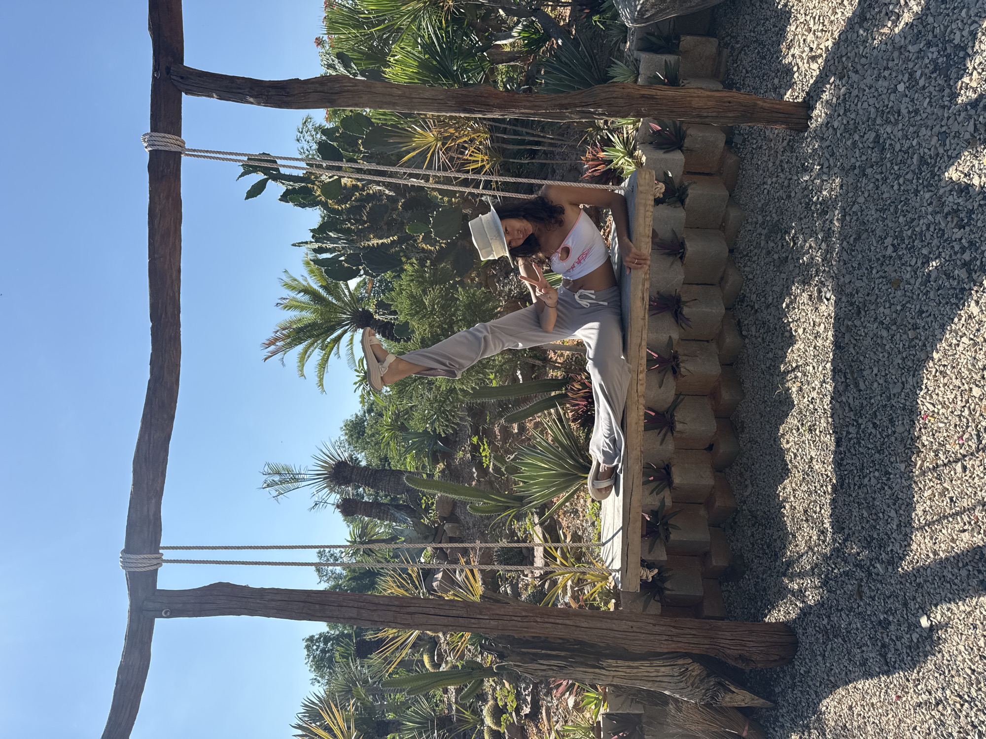 In Mũi Né, Vietnam, a young woman with dark curly hair and a white sun hat reclines on a wooden swing suspended from a rustic wooden frame. She wears a white tube top with a pink trim and loose grey pants, with one leg extended upwards and the other bent. Her right hand is raised in a peace sign. The swing is situated in a garden filled with various succulents and cacti, under a clear blue sky. The ground is covered in small grey stones, and the scene is bathed in bright sunlight, casting shadows from the swing's frame.