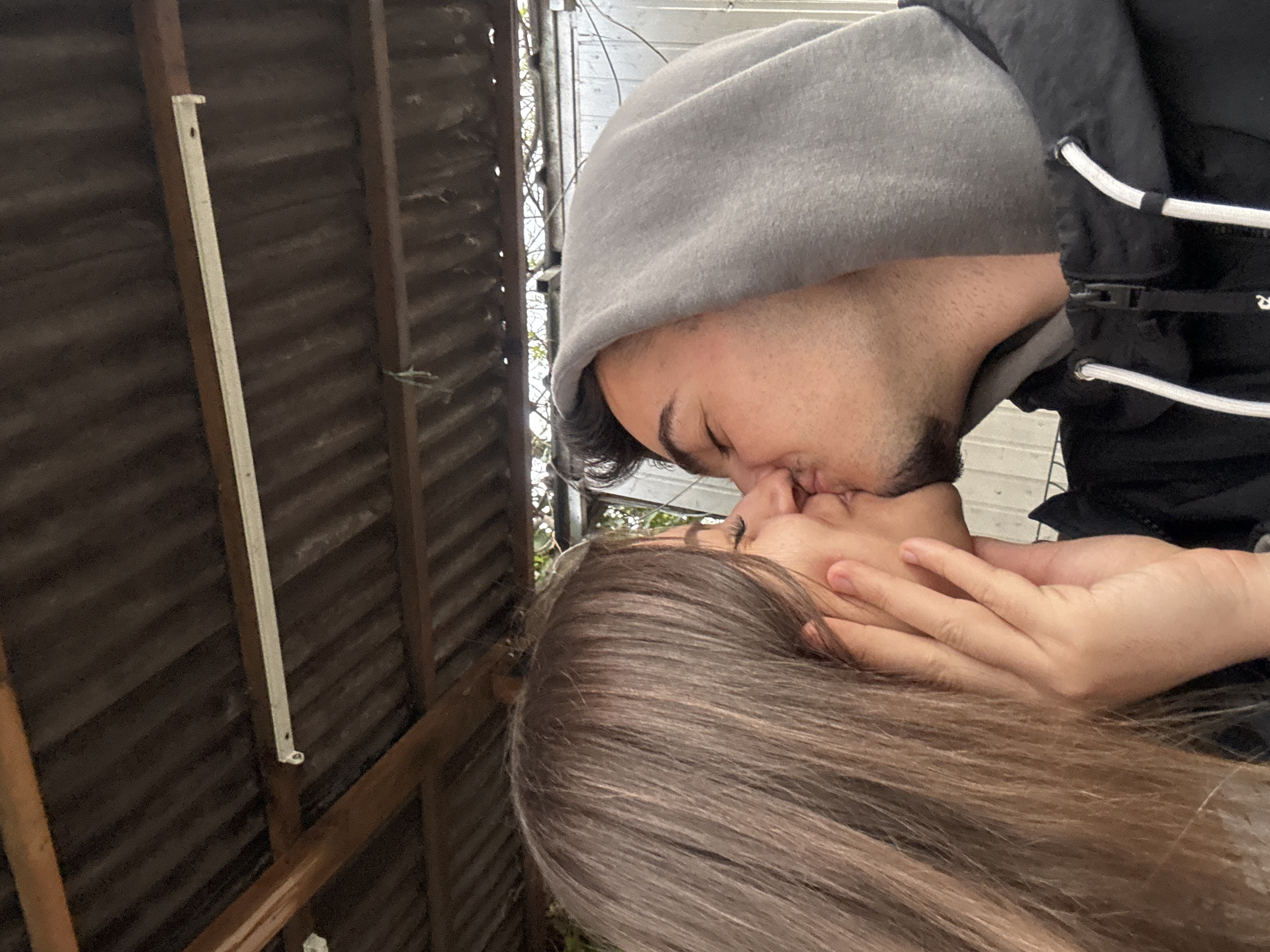 The image captures an intimate moment between a man and a woman kissing, with the photo taken from a slightly tilted perspective. The man is seen from the upper right, wearing a grey hooded sweatshirt with the hood up and a black jacket featuring white drawstrings. He has dark hair and light stubble on his face, and his eyes are closed as he kisses the woman.
The woman, with long, straight brown hair cascading around her face, is positioned below and to the left of the man. Her hand is tenderly placed on the side of his face/neck as they share the kiss. She wears glasses, partially visible through her hair. Both individuals appear engrossed in the embrace.
The background consists of dark, possibly corrugated metal siding or fencing, supported by visible brown wooden beams. A narrow white vertical strip, possibly a trim or fixture, is attached to one of the wooden beams on the left. Faint traces of sparse vegetation or branches are visible in the upper background against a lighter, possibly overcast, sky. The lighting is soft and diffused. The scene suggests a private, outdoor setting, possibly a backyard or an area next to an outbuilding in Sofia, Bulgaria. There is no visible text in the image.