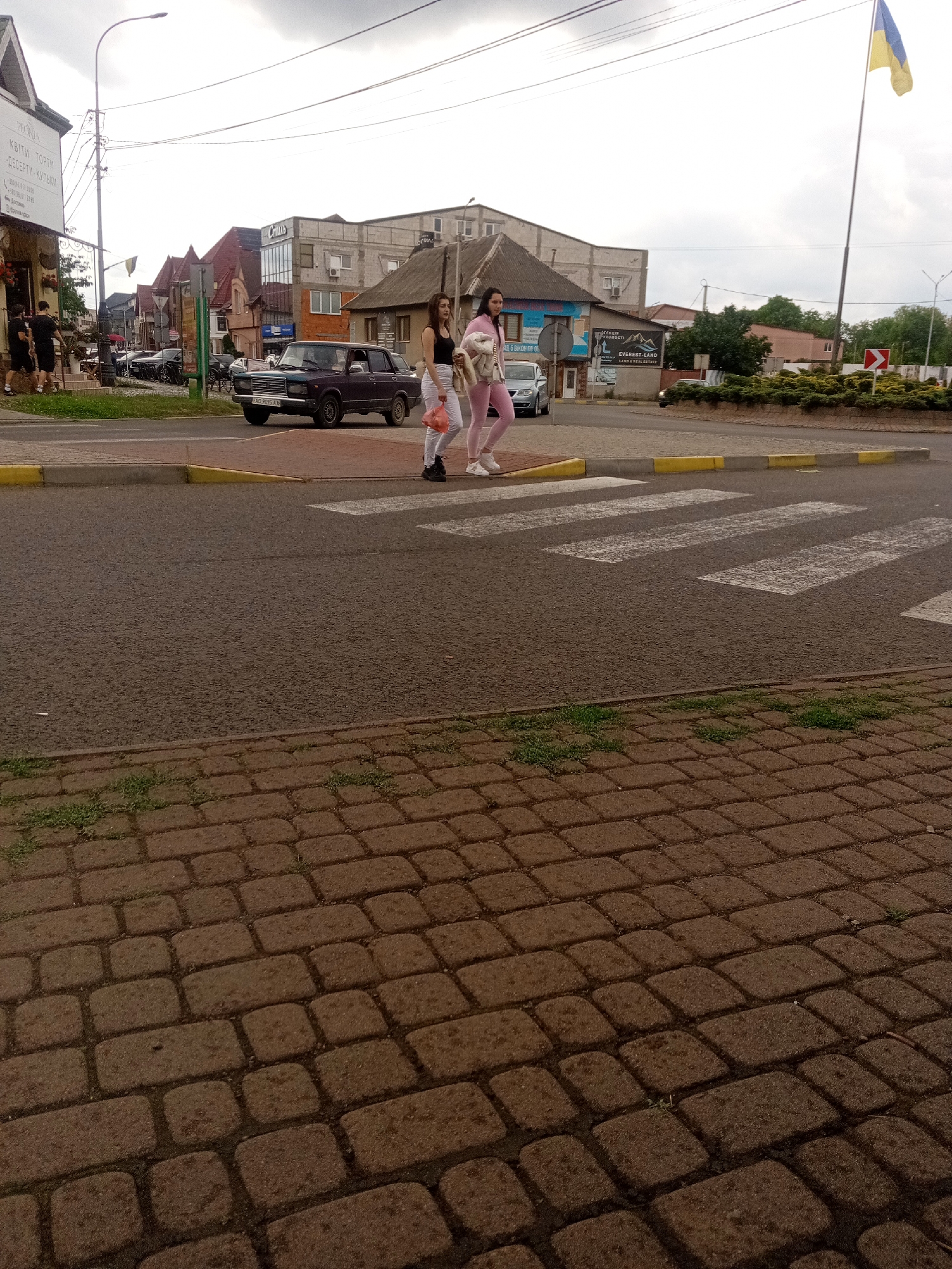 A scene unfolds on a street in Tiachiv, Ukraine. A crosswalk dominates the foreground, with two young women approaching it from the right. The woman on the left wears a black tank top and white pants, while the woman on the right sports a pink athletic outfit. The latter carries a light-colored bundle, possibly clothing or a bag.
Behind the women, a dark sedan with license plate "AA 5095 AK" is stopped on the road, likely yielding to pedestrians. The street is lined with buildings, some with distinctive pitched roofs, suggesting a mix of residential and commercial establishments. A prominent Ukrainian flag flies from a tall pole to the right of the frame, a poignant reminder of the country's national identity. The sky overhead is overcast, casting a muted light on the urban landscape. The sidewalk in the foreground is paved with reddish-brown interlocking bricks.