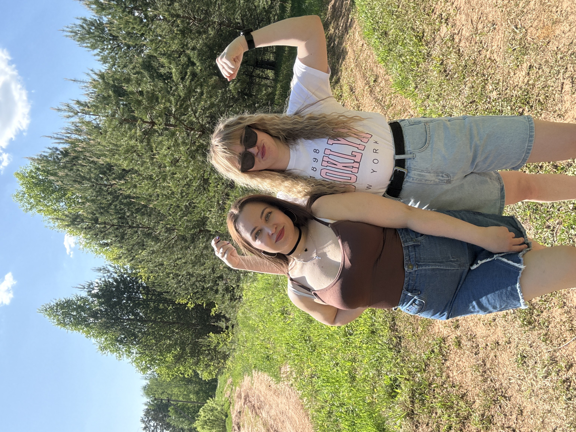 Two young women are posing for a photograph outdoors in a natural setting. The woman on top has blonde, curly hair, sunglasses, and a white t-shirt with "BROOKLYN NEW YORK" printed on it. She is wearing light-wash denim shorts with a black belt. Her right arm is raised in a flexing pose. The woman on the bottom has brown hair, a brown tank top, and dark-wash denim shorts with a frayed hem. She is also posing with one arm raised. They are standing on a grassy area beside a dirt path. In the background, there are numerous trees, and the sky is blue with a few white clouds. The image was taken in a natural environment, however, I am unable to confirm the location in the city of Марковский, Russia, based on the information in the image.