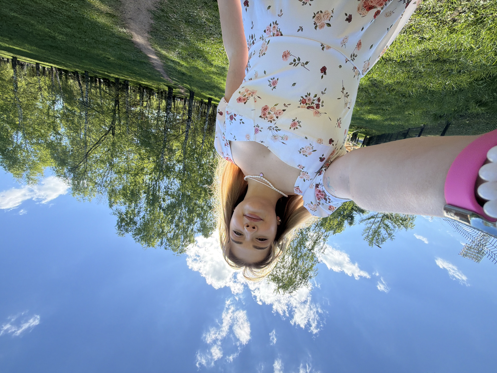 The image, rotated 180 degrees, depicts a young woman in a white dress with a floral pattern. She wears a pearl necklace. Her face is visible, and she appears to be outdoors. The background includes a clear blue sky with scattered white clouds, as well as green trees and grass. The location appears to be a park. The image might be taken in Naberezhnye Chelny, Russia.