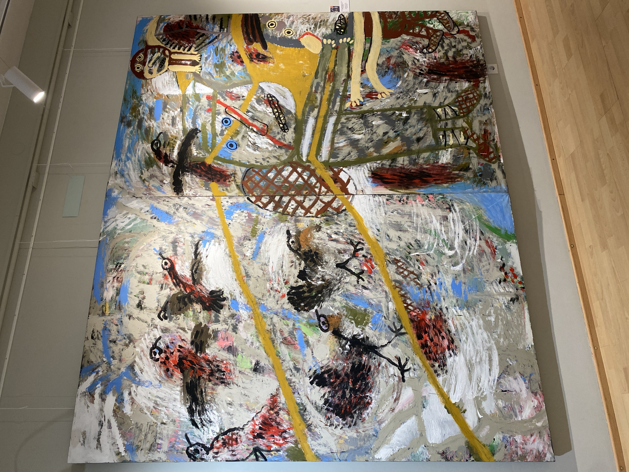 The image depicts a large, diptych painting displayed in a gallery setting. The painting is abstract and features bold, energetic brushstrokes in a variety of colors including white, grey, blue, yellow, red, and brown. Several stylized bird-like figures are prominent throughout the work, rendered with simple shapes and dark outlines. The right panel of the diptych also contains figures that resemble stylized humanoids with large, round eyes and outstretched limbs, painted in yellow, brown, and red. Yellow diagonal lines are a recurring motif in the composition. The painting hangs on a light grey wall, illuminated by track lighting. A light-colored wooden floor is visible in the foreground. A small white label with text is affixed to the right side of the painting, partially visible. The overall impression is one of a dynamic and expressive piece of contemporary art. The provided image does not contain information indicating its specific location in the city of Petrozavodsk, Russia.