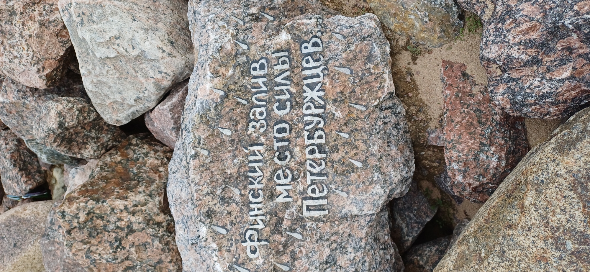 The image shows a cluster of granite stones. On one of the stones, there is an inscription in white paint in Russian, which reads: "Финский залив место силы Петербуржцев," translating to "Gulf of Finland, place of power of Petersburgers". The location is likely a coastal area in or near Saint Petersburg, Russia. The image suggests a natural setting with human interaction (the inscription on the rock).
