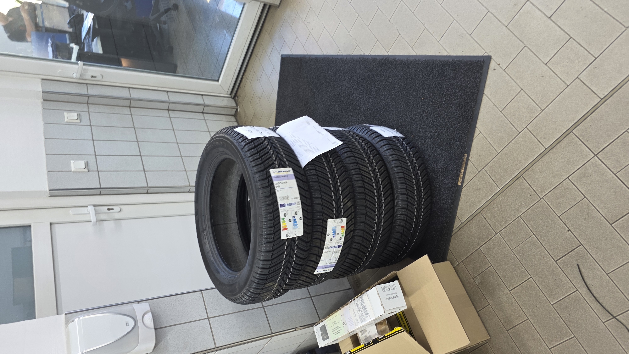 A stack of four black tires sits on a dark grey mat on a light beige tile floor. Each tire has a white sticker with European Union energy efficiency labeling and other tire specifications. To the left of the tires is an open cardboard box containing some packaging material and a white paper. Behind the tires is a wall tiled with small, light grey square tiles. To the right of the tiled wall is a large glass door with white framing. In the reflection of the glass door, a person can be seen standing at a desk with a computer monitor. A white soap dispenser is mounted on the tiled wall to the left of the door. The overall scene appears to be in an indoor service or retail environment.