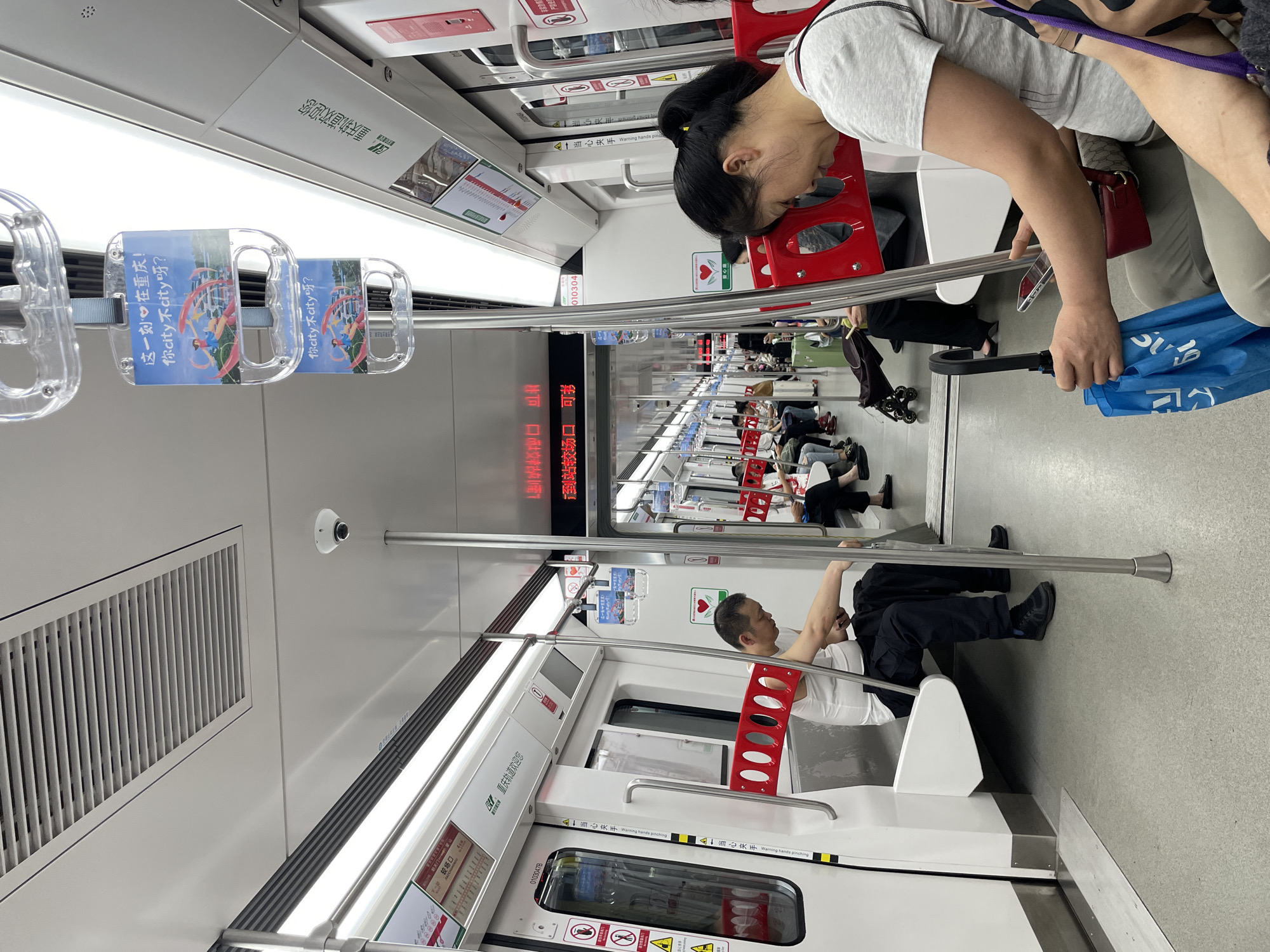 This is an indoor shot of the interior of a subway car in the Yuzhong District of Chongqing, China. Several individuals are present: one woman stands at the top right, appearing tired as she rests her head on a vertical support pole. Another man is seated near the bottom right of the frame, dressed in what appears to be a white shirt and dark pants. Multiple other passengers are seated further down the subway car, adding to the scene's ambiance.
Various objects and features are discernible, including clear plastic handles with advertisements on them, red vertical support poles, and electronic displays showing station names. The interior design of the subway car is predominantly white and gray, contributing to a modern aesthetic.
Overall, the scene captures a typical moment in the daily commute within Chongqing's public transportation system.