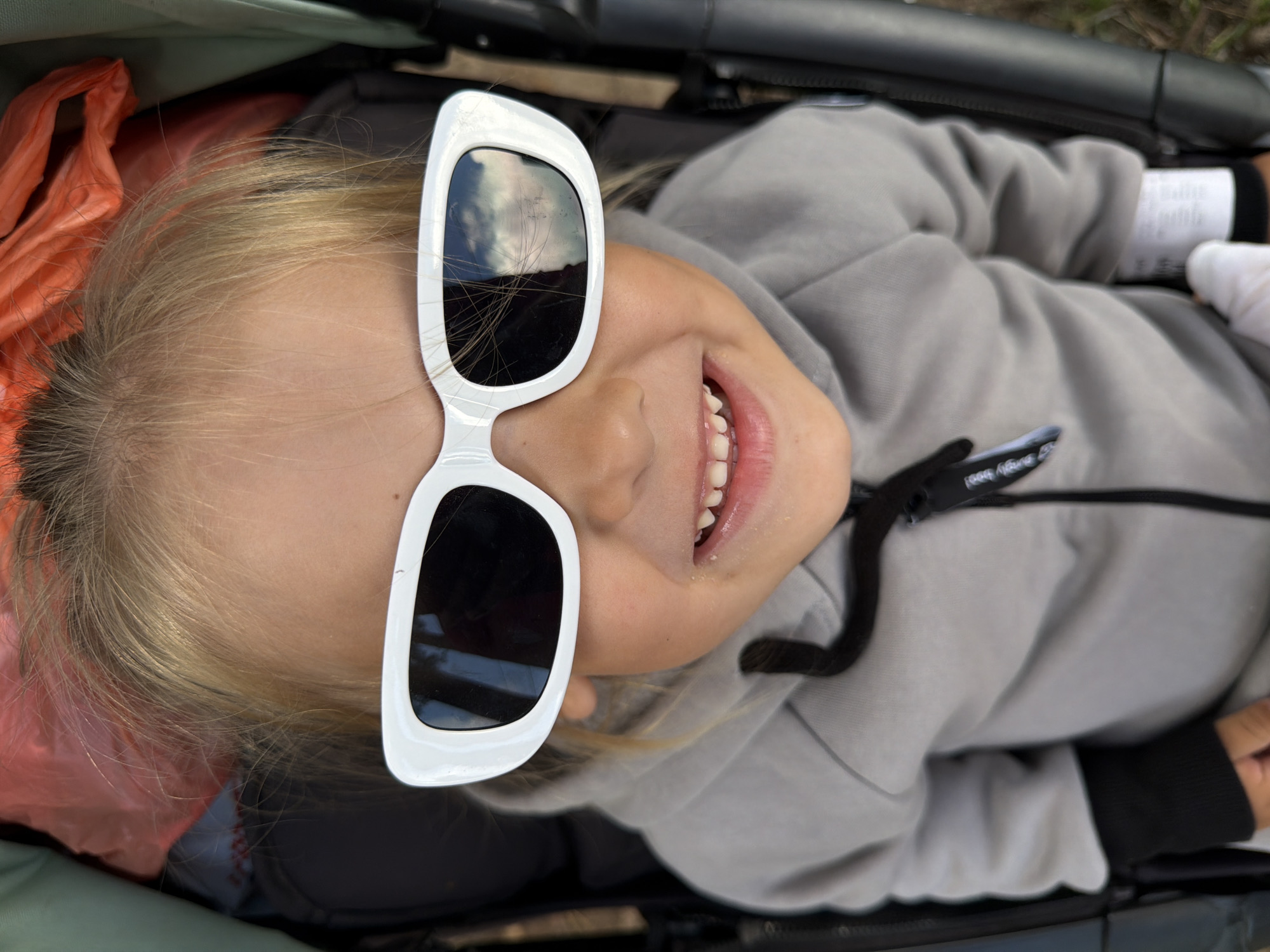 A close-up, high-angle shot captures a young blonde girl wearing oversized white rectangular sunglasses with black lenses. She is smiling and looking directly at the camera, revealing her teeth. The girl is wearing a grey hooded jacket with a black zipper pull and black cuffs. The background is blurred and appears to be outdoors, possibly in a park or natural setting, with hints of greenery and brown earthy tones. The bright orange material behind the girl's head suggests she may be in a stroller or some form of seating with a canopy. The overall impression is a candid moment of a child enjoying an outing. The location in Chaikovsky, Russia is not ascertainable from the visual information provided in the image.