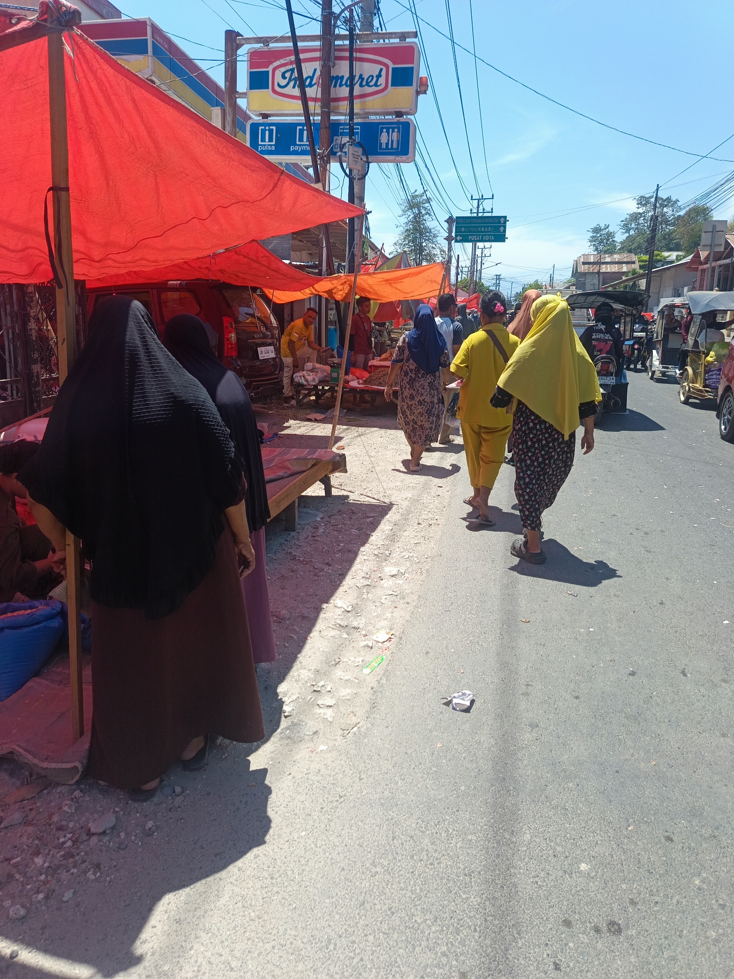 The image shows a street scene in Gorontalo, Indonesia, on a sunny day. In the foreground, two women wearing black and brown hijabs are standing under a red awning, facing away from the camera. To their right, three women are walking down the street. The woman on the far right wears a yellow hijab and a black and floral patterned skirt. The woman in the middle wears a yellow top and pants. The woman on the left wears a blue hijab and a floral patterned dress. In the background, there are several buildings, including an Indomaret store, and various vehicles, including motorcycles and a tuk-tuk. There are also several market stalls with vendors and goods. The street is paved and appears to be busy with people and traffic.