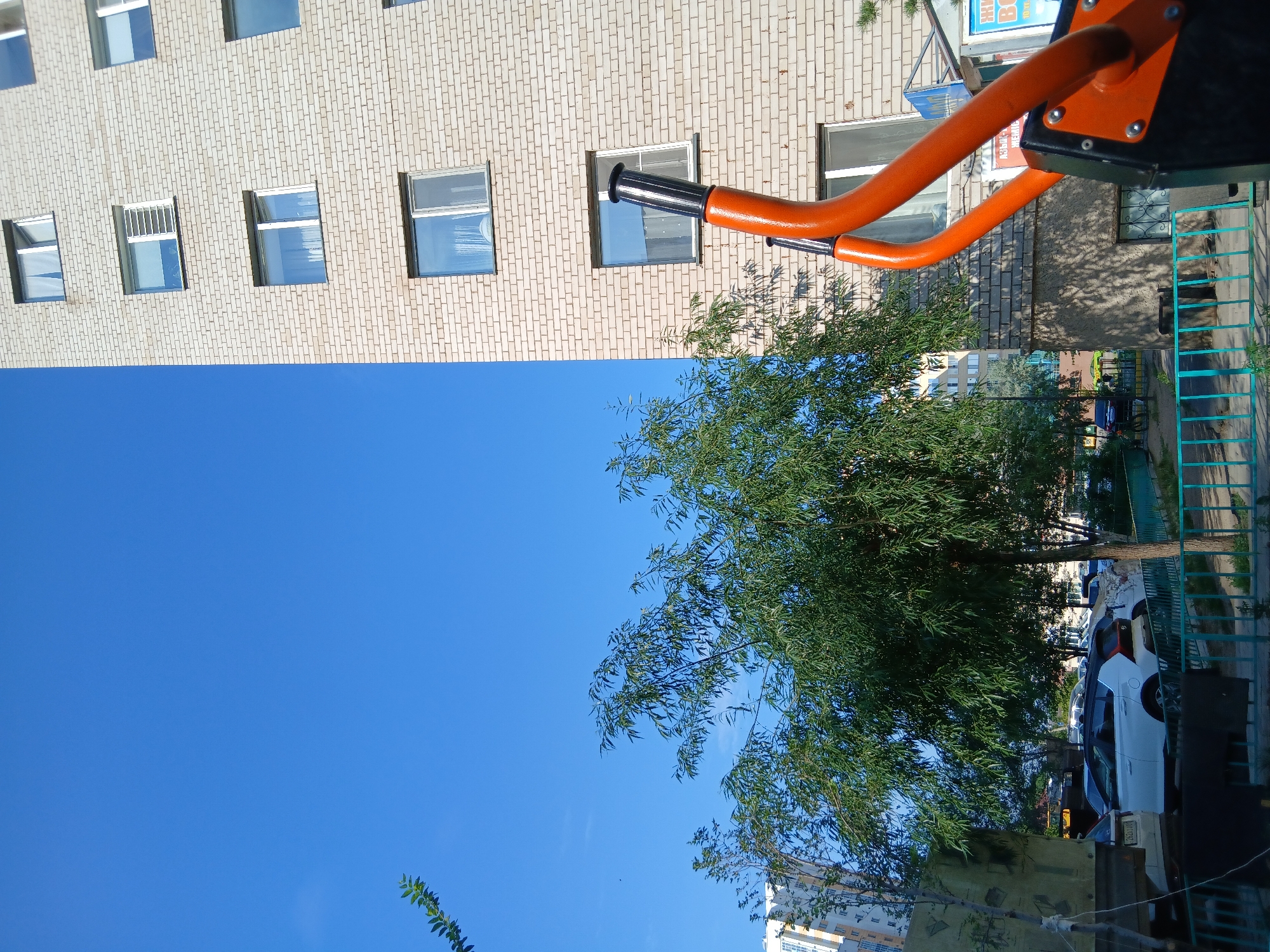 The image shows an outdoor scene with a brick building and a tree. On the top right, there is an orange and black exercise machine, likely part of an outdoor gym. The building has several windows, some with white frames and some with darker frames. The tree is green and leafy, with its branches extending towards the sky. The sky is clear and blue, suggesting a sunny day. In the background, there are cars parked on the street and other buildings. A turquoise metal fence is visible on the right side of the image. The scene appears to be a residential area or a park. There are no people visible in the image. There is some text on a sign on the building in the upper right, but it is too blurry to read. The image is taken from a low angle, looking up towards the building and sky. The overall mood is calm and peaceful, with the natural elements of the tree and sky contrasting with the man-made structures. The location context provided is Astana, Kazakhstan, which helps to understand the architectural style and climate.