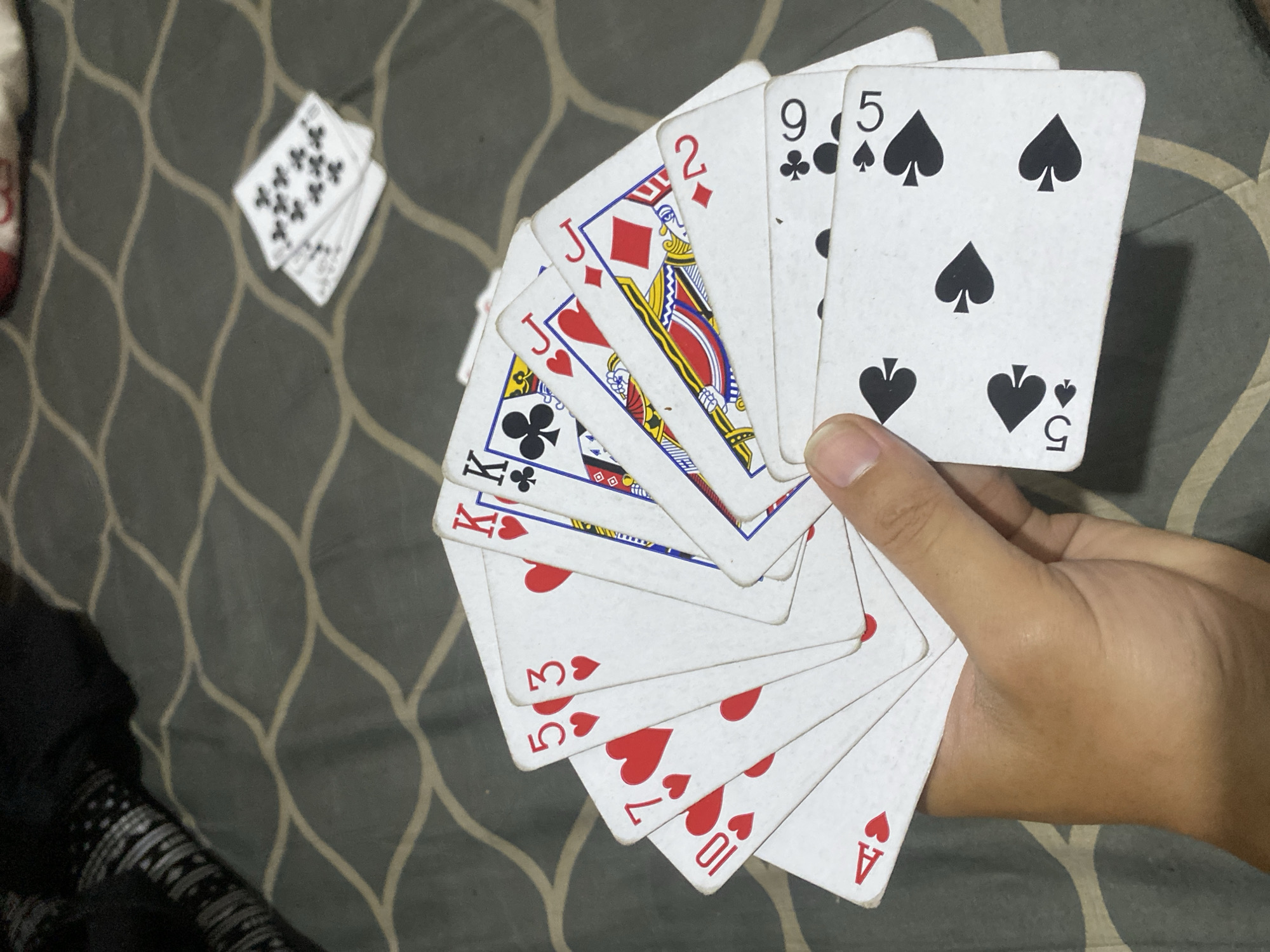 A hand holds a fan of playing cards, which include the Ace, 10, 7, 5, and 3 of Hearts, the King of Clubs, Jack of Hearts, 2 of Diamonds, 9 of Clubs, and 5 of Spades. A partial stack of cards with clubs is visible in the background. The scene is likely indoors, the city of Bandung City, Indonesia, given the prompt. The hand is tanned. The background is a textured, patterned surface.