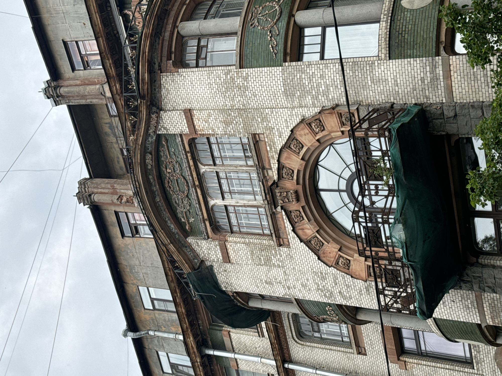 The image shows the facade of a historic building in Saint Petersburg, Russia, likely an apartment building, with an overcast sky. The architecture features a mix of materials and styles, including white brickwork, green tiled sections with decorative reliefs, and reddish-brown stonework around arched windows and balconies. One balcony is covered by a green tarp. Several windows are visible, some with decorative frames. There are electrical wires crossing the sky in the upper portion of the image. The building appears aged and somewhat weathered, suggesting a long history. The lower part of the building shows a darker, rougher stonework foundation. Green foliage is visible at the very bottom of the frame.