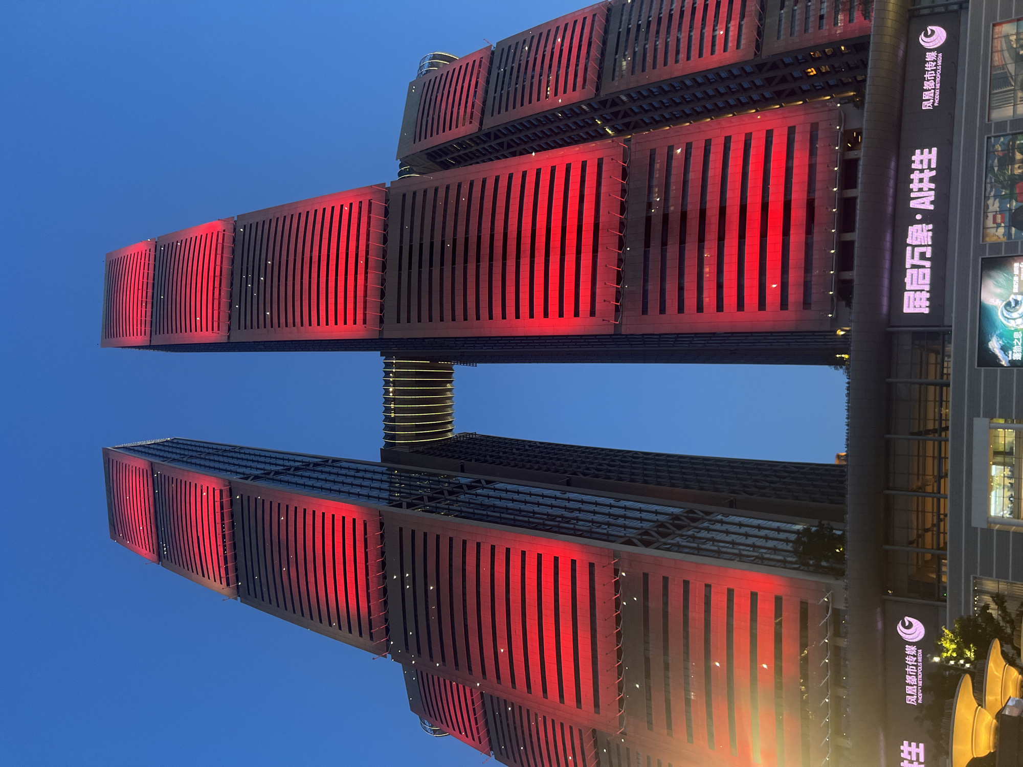 This is a vertical, low-angle outdoor shot of the Fenghuang Building, also known as Phoenix Building, located in the Yuzhong District of Chongqing, China. The two dominant towers of the building are illuminated with bright red lights, creating striking patterns across their facades. The towers are connected by a skybridge which is also illuminated with horizontal yellow lights. The building's architecture features a repeating pattern of horizontal dark lines that contrast with the red illumination. The sky is a deep blue, indicating it is dusk or early evening. At the base of the towers, a large electronic billboard displays text in Chinese characters and an English translation, "PHOENIX METROPOLIS MEDIA," alongside a circular logo. There are no people visible in the image. The scene depicts a modern urban architectural landmark at night.