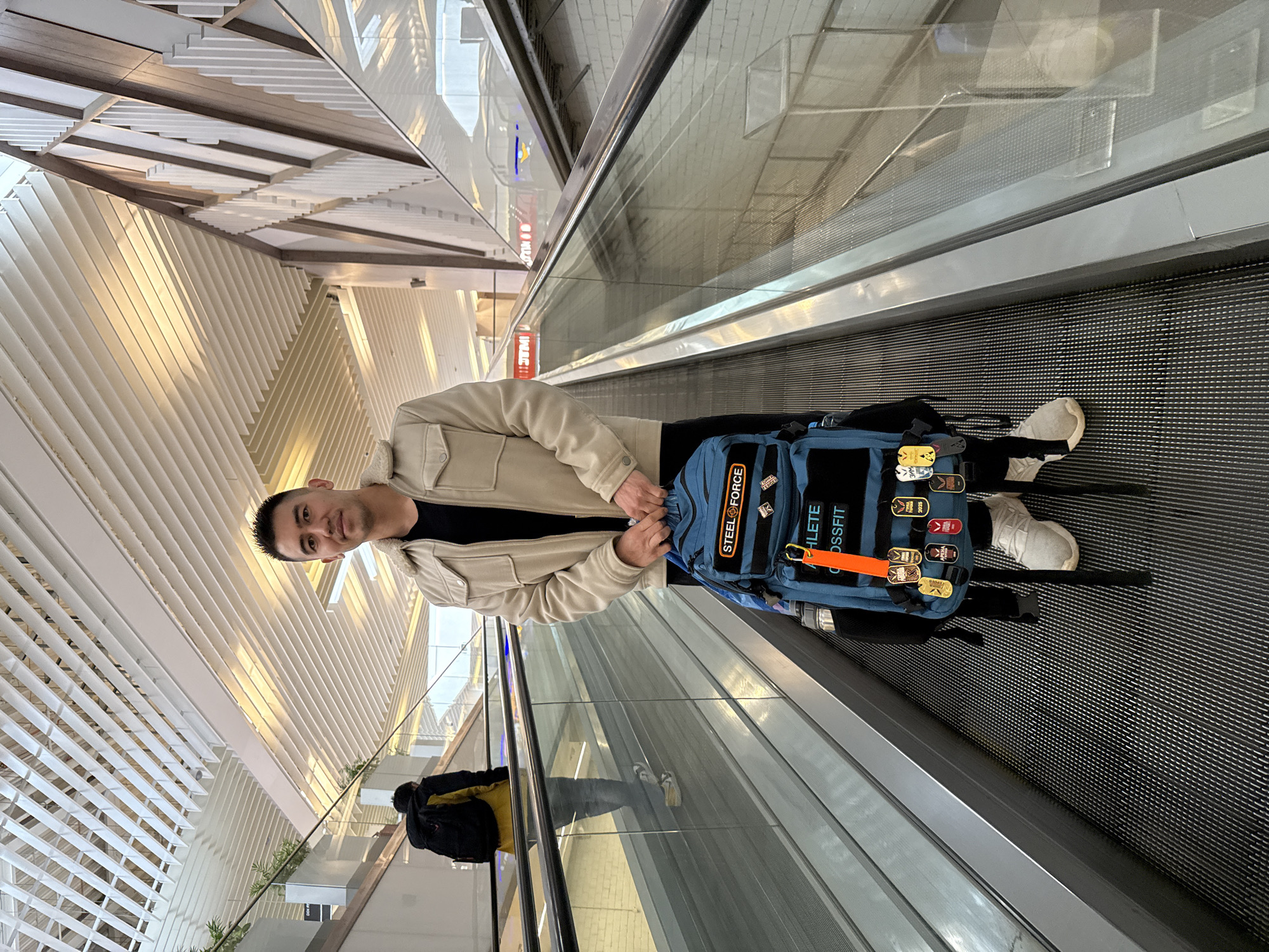 The image shows a man riding a moving walkway, likely at an airport, in Kazan, Russia. The man is wearing a beige jacket over a black shirt, and he has a blue backpack with several patches on it, including the words "STEEL FORCE" and "ATHLETE CROSSFIT". He is also wearing white sneakers. The moving walkway is surrounded by glass barriers and has a black, ribbed surface. The background features a modern architectural design with white and beige structures. A person with a yellow and black backpack can be seen riding the escalator below the man.