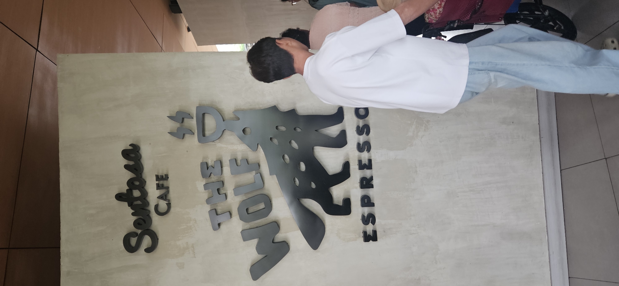 The image shows a sign for "Sentosa CAFE THE WOLF ESPRESSO" mounted on a textured concrete wall. The sign features black, three-dimensional lettering and a silhouette of a wolf. To the right of the sign, a person wearing a white t-shirt and light blue pants is partially visible, facing away from the camera and appearing to be walking. Another person is partially visible behind them. The background includes a modern ceiling structure and a glimpse of a building's exterior. The scene is consistent with an outdoor area of a cafe in a developed urban setting.