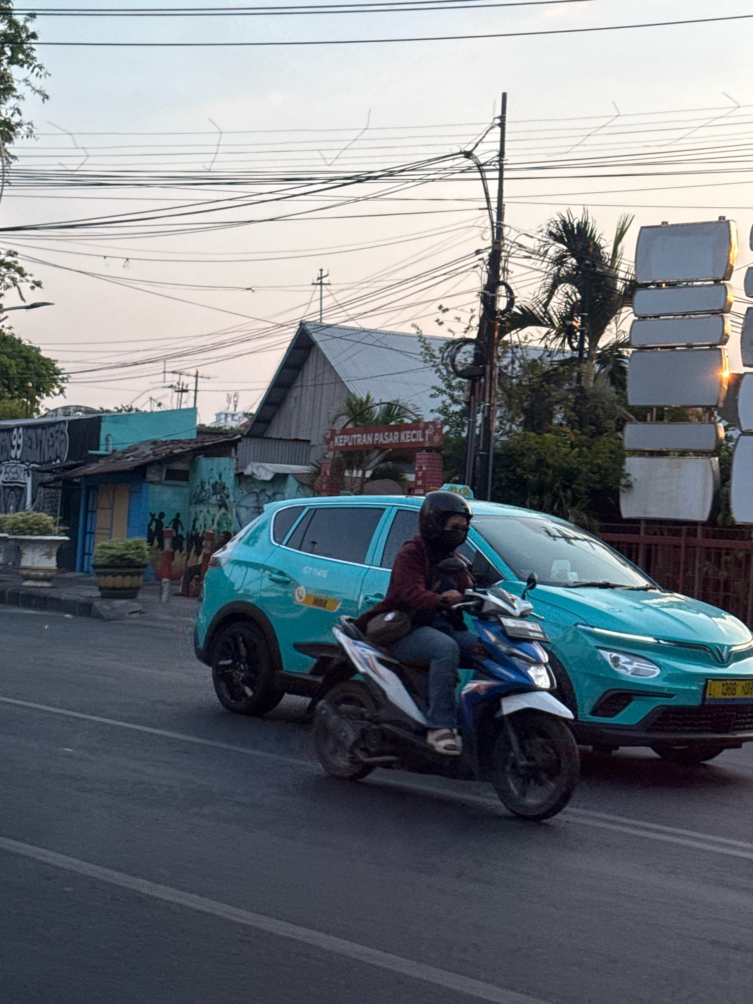 The image shows a street scene in Surabaya, Indonesia, with a turquoise car and a motorcycle in motion. The car, which appears to be a taxi, has a license plate starting with "L", common for East Java, Indonesia, and the text "KEPUTRAN PASAR KECIL" on a sign. The motorcycle is being ridden by a person wearing a helmet and mask. The background features several buildings, including a blue building with murals and some signage, along with utility poles and overhead wires. The overall lighting suggests it's either early morning or late afternoon.