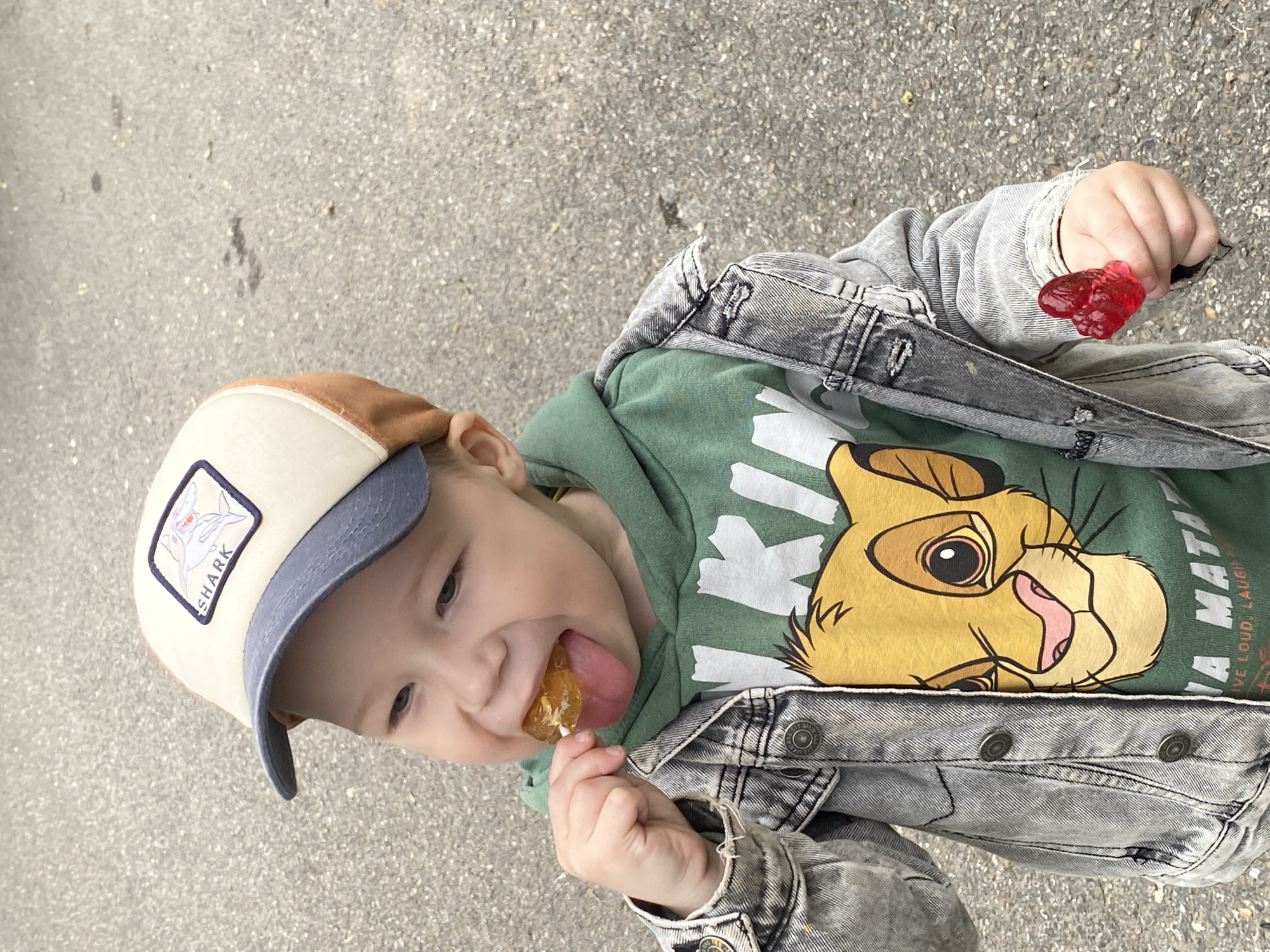 A young child wearing a baseball cap, a green t-shirt with a lion cartoon, and a denim jacket is eating a lollipop. The child is holding a second, heart-shaped candy in their other hand. The child is standing on asphalt. This scene does not provide enough information to determine if it's located in Sochi, Russia.