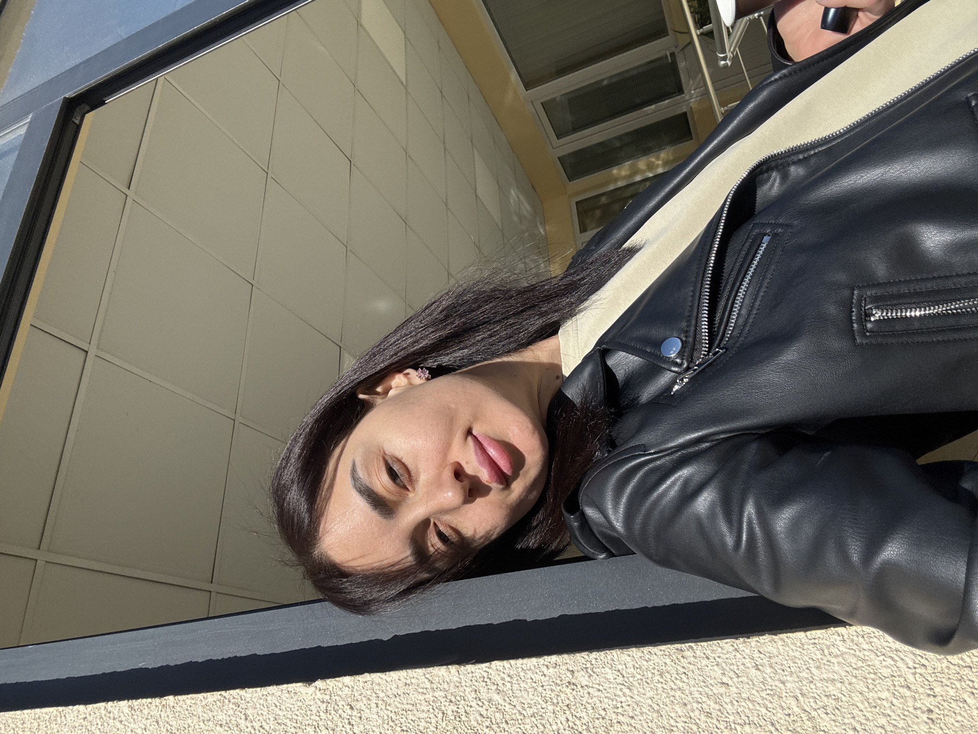 The image depicts a person with dark hair and a black leather jacket leaning against a structure with a grid-like pattern on its surface. The person is looking at the camera. The location is potentially in Tbilisi, Georgia. The environment appears to be outdoors during daylight.