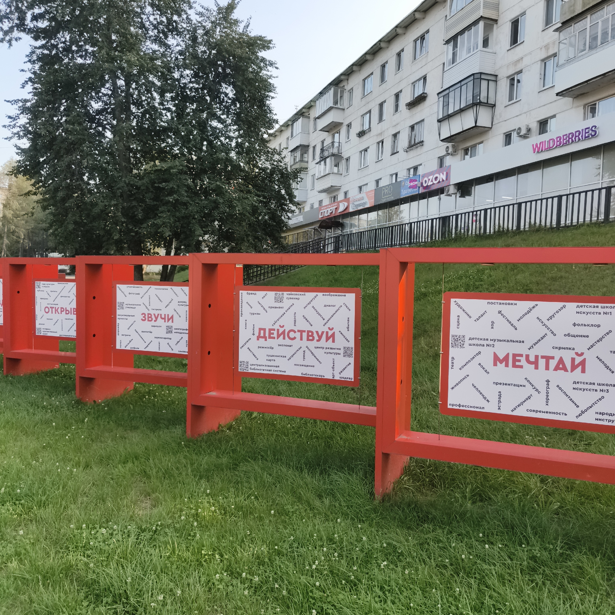 The image shows a red metal fence with several white rectangular signs attached to it, displaying Russian text and QR codes. The fence is set up on a grassy area in front of a modern apartment building. The building has multiple floors and balconies, with shops on the ground level. Some of the shop signs visible include "WILDBERRIES," "OZON," and "SPORT." A large tree with dense green foliage is on the left side of the frame, obscuring part of the building. The signs on the fence appear to be part of an outdoor exhibition or information display. The text on the signs includes words such as "ДЕЙСТВУЙ" (ACT), "ЗВУЧИ" (SOUND), and "МЕЧТАЙ" (DREAM), along with various related terms and possibly event information or educational themes. The scene is well-lit, suggesting it is daytime, and the overall impression is of a public space in an urban environment.
