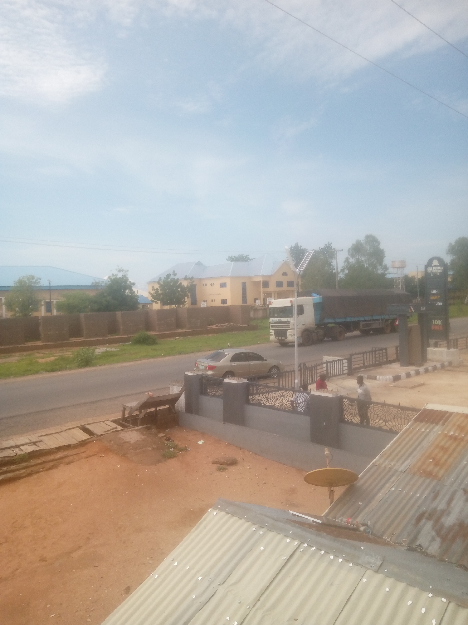 This image depicts an outdoor street scene in Jalingo, Nigeria, captured from an elevated perspective during daytime under a bright, partly cloudy sky.
In the immediate foreground, a light-colored corrugated metal roof with visible fasteners occupies the lower right quadrant. A small, circular, gold-colored satellite dish on a pole is mounted on this roof. Adjacent to the roof is a patch of reddish-brown dirt, leading to a low wall topped with dark-colored, intricately patterned metalwork featuring a radial or web design.
Three adult males are visible standing behind this decorative wall, near the street level. One man wears a red shirt, another a light-colored shirt, and the third is partially obscured.
An asphalt road runs horizontally through the midground. A light-colored sedan, possibly beige or gold, is parked on the left side of the road, behind the decorative wall. A large white DAF-brand truck, with its open cargo bed covered by a dark blue tarp, is traversing the road from left to right. The cargo beneath the tarp appears dark and bulky.
On the right side of the road, a vertical commercial sign displays "SULEIMAN OVERTURE LTD," "MINI MARKET," "GAS," and "JPK DIMS." Behind this sign, an elevated water tank on a metal stand is visible. Faint power lines span across the sky. On the left side of the road, a wooden bench or makeshift table rests on a dirt verge.
In the background, several buildings are present. On the far left, a large structure with a prominent blue corrugated roof is visible. Towards the center-right, a multi-story building with a light yellow facade and dark windows, topped with a grey or blue roof section, stands out. Additional buildings and numerous green trees and shrubs are scattered throughout the background, indicating a vegetated environment. A low, brown-colored wall runs parallel to the road, separating it from the properties in the distance. The ground in the foreground and along the verges is primarily reddish-brown dirt.