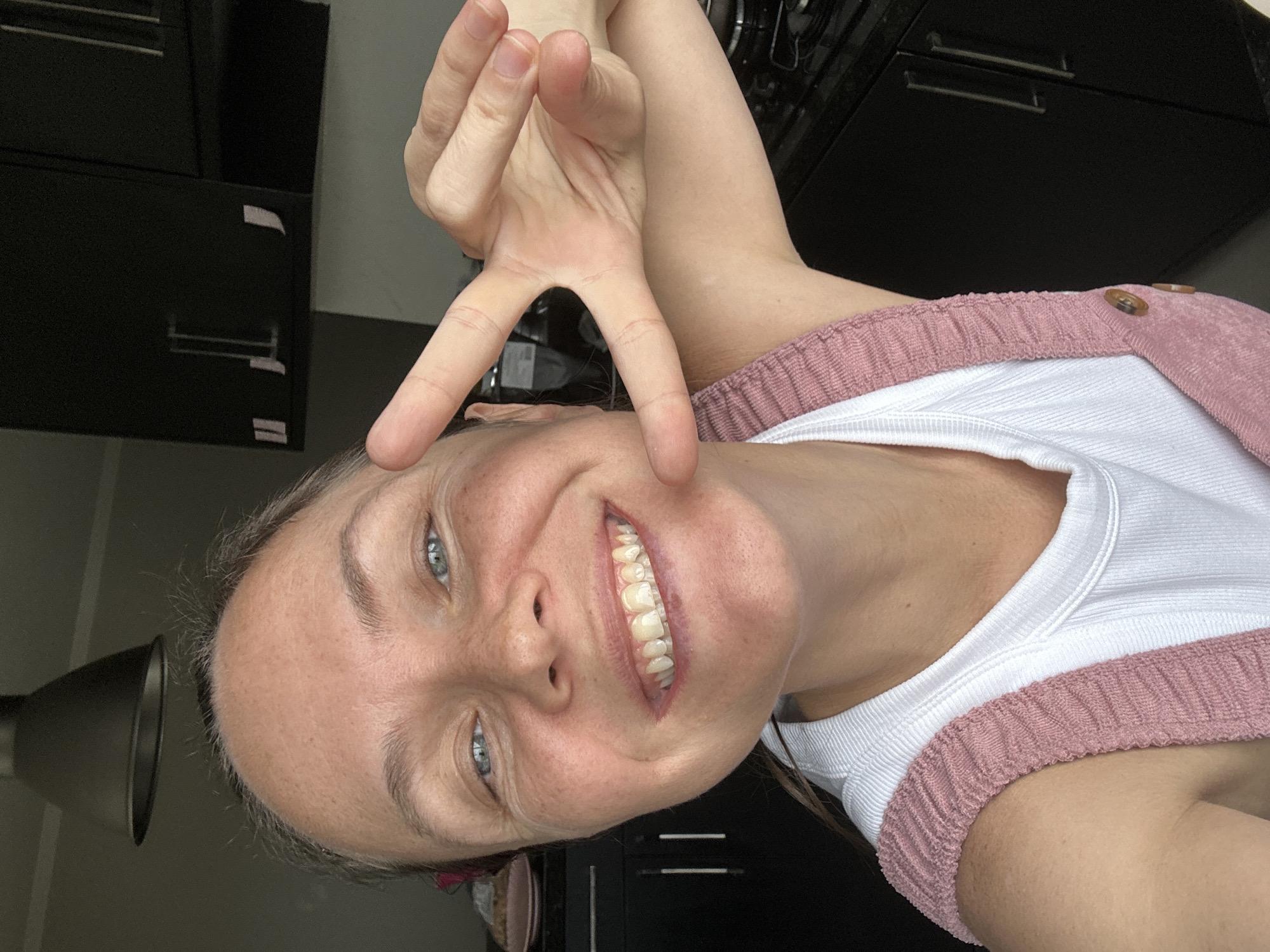 A woman is taking a selfie in what appears to be a kitchen. She is smiling with her teeth showing and making a peace sign with her right hand. She is wearing a white tank top and pink overalls. In the background, there are dark cabinets, a stove, and a ceiling light fixture. The image quality suggests it was taken with a mobile phone camera. The lighting is natural, coming from the side. The woman's skin has freckles and is slightly tanned. The environment suggests a residential setting. Florianópolis is a city in Brazil known for its beaches and natural beauty.