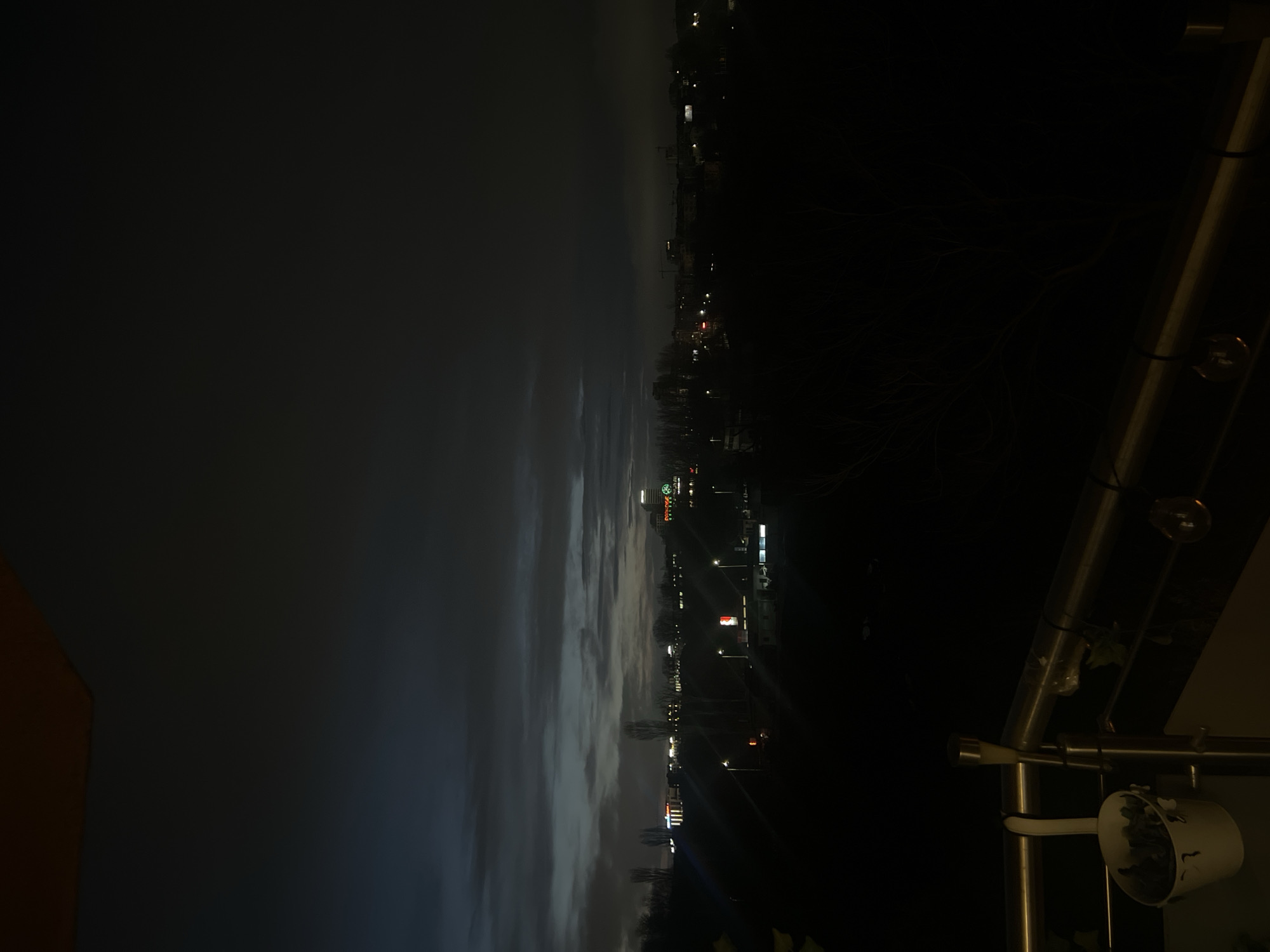 The image depicts a night scene in Sofia, Bulgaria. The view is from a high vantage point, likely a balcony, looking over a part of the city. In the distance, city lights illuminate buildings and streets, with a few prominent commercial signs visible. The sky is dark and cloudy, with some lighter areas near the horizon. In the foreground, the balcony railing and some external lighting fixtures are visible.