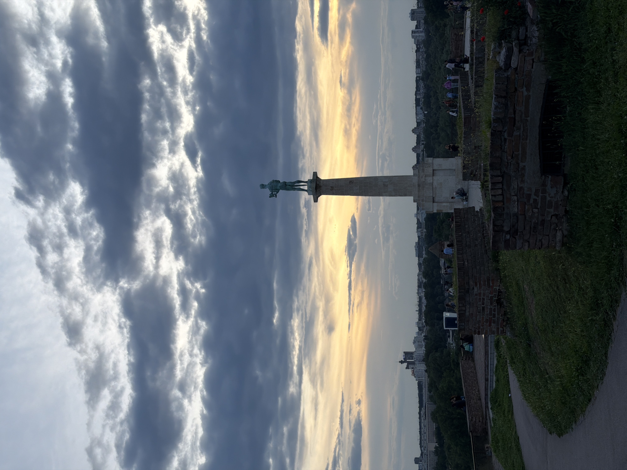 The image shows the Victor Monument in Belgrade, Serbia, with the statue facing left. The sky behind the monument is filled with striated clouds, dark on the left and bright on the right, suggesting the sun is low on the horizon. To the right of the monument, the view extends over the city, with buildings visible in the distance, partially obscured by foliage. The foreground shows a brick wall, some greenery, and a pathway where people are situated.