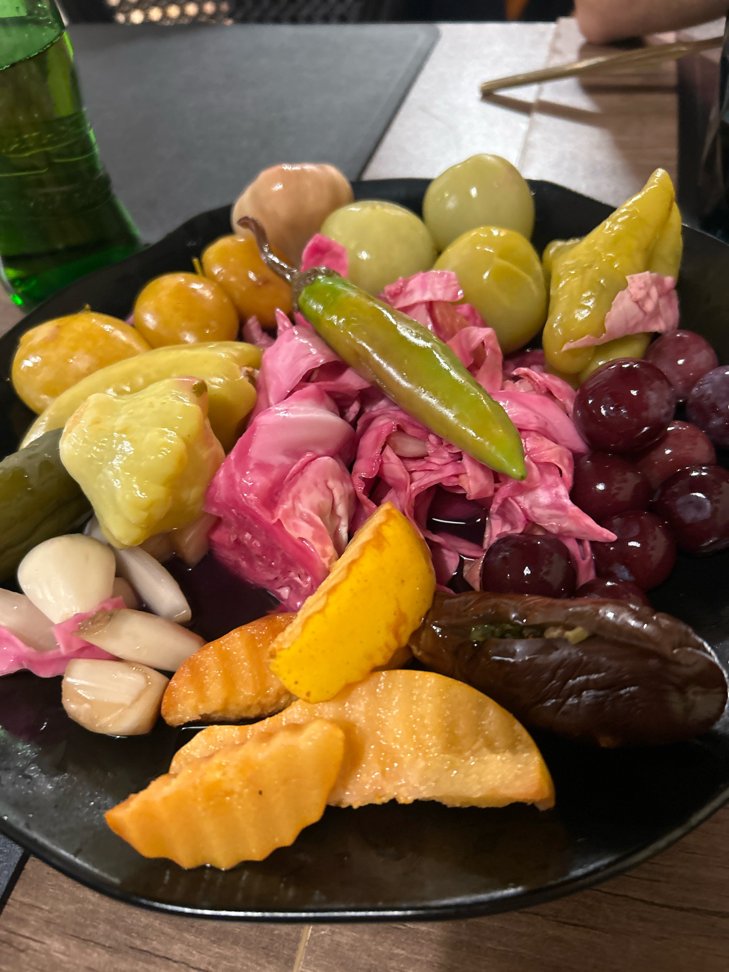 A platter of assorted pickled vegetables and fruits is presented on a dark plate. The platter contains green pickles, small yellow pickled peppers, pickled tomatoes, what appears to be pickled cabbage with a pinkish hue, a green chili pepper, dark red grapes, pickled garlic cloves, pickled peach slices, and a stuffed eggplant. A green glass bottle is visible to the left of the platter. The platter is placed on a wooden table. The background shows a blurred indoor setting with some decorative elements, possibly in Sabail Raion, Azerbaijan. There are no visible people in the foreground.