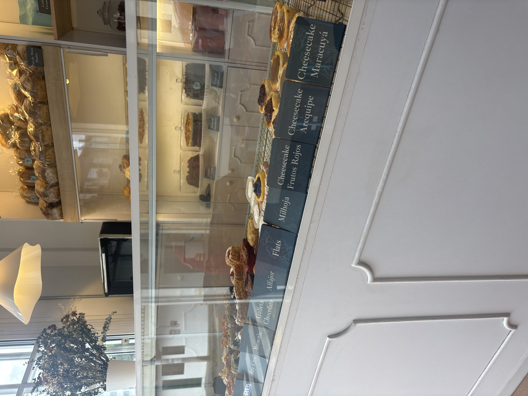 This image depicts a bakery display case in Guarne, Colombia. The case holds a variety of pastries and desserts, including different flavors of cheesecake (maracuyá, arequipe, frutos rojos), milhoja, flats, alfajor, chocolate cookies, brownies, and rey croi. The image also shows a vase with dried flowers, a lamp, and window light from the outside. "Pan de Masa Madre" is visible, along with "Cafe Gran."