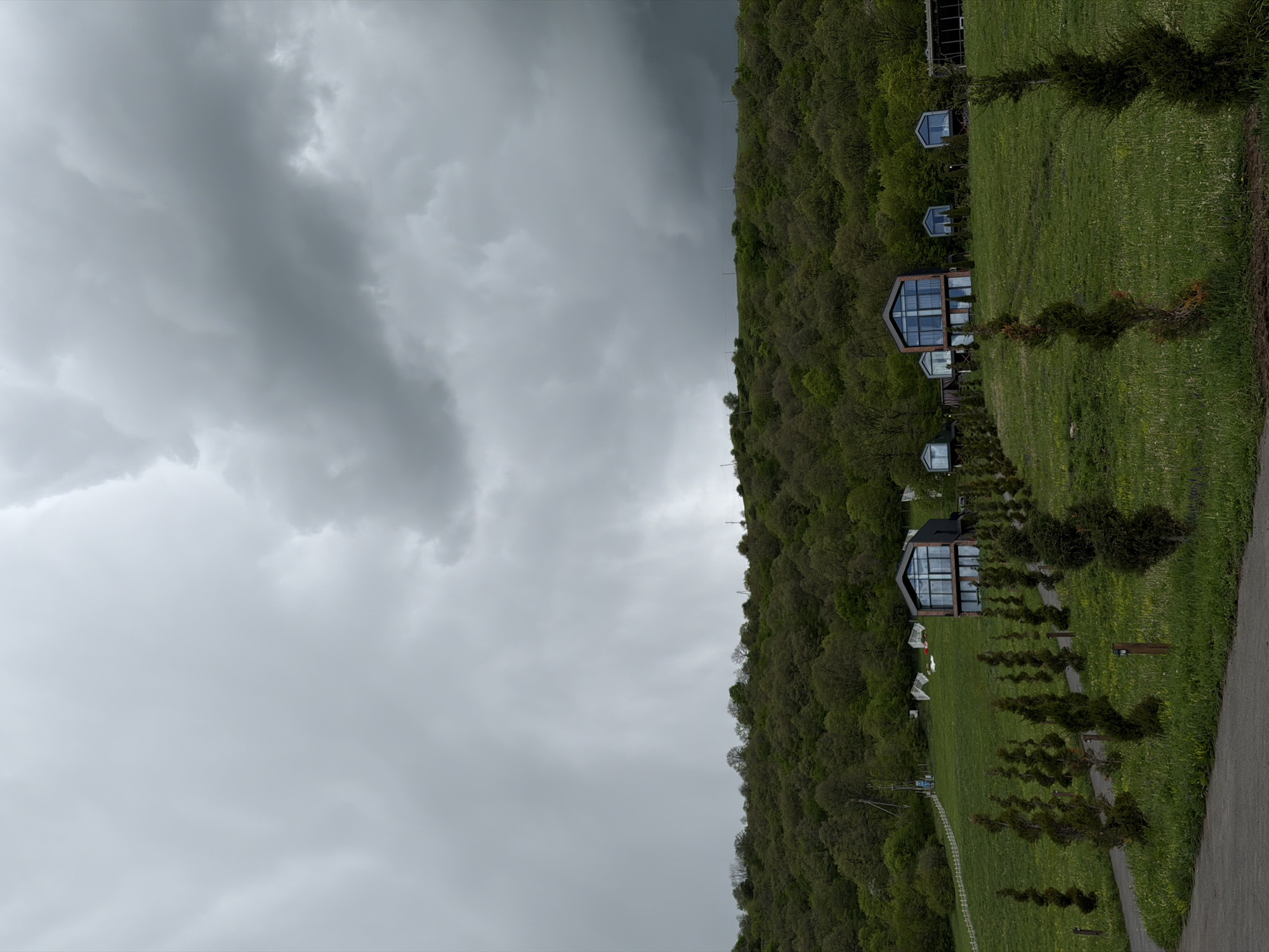 The image depicts a series of modern, chalet-style buildings nestled on a hillside, likely in a rural setting. The structures have a distinctive architectural design featuring large windows. The surrounding terrain is characterized by lush green grass, trees, and foliage. Overhead, the sky is overcast with heavy, gray clouds, suggesting a potentially stormy or dreary weather condition. A path or paved area is visible in the foreground. This scene is most likely located near Dsegh, Armenia, given the context provided.