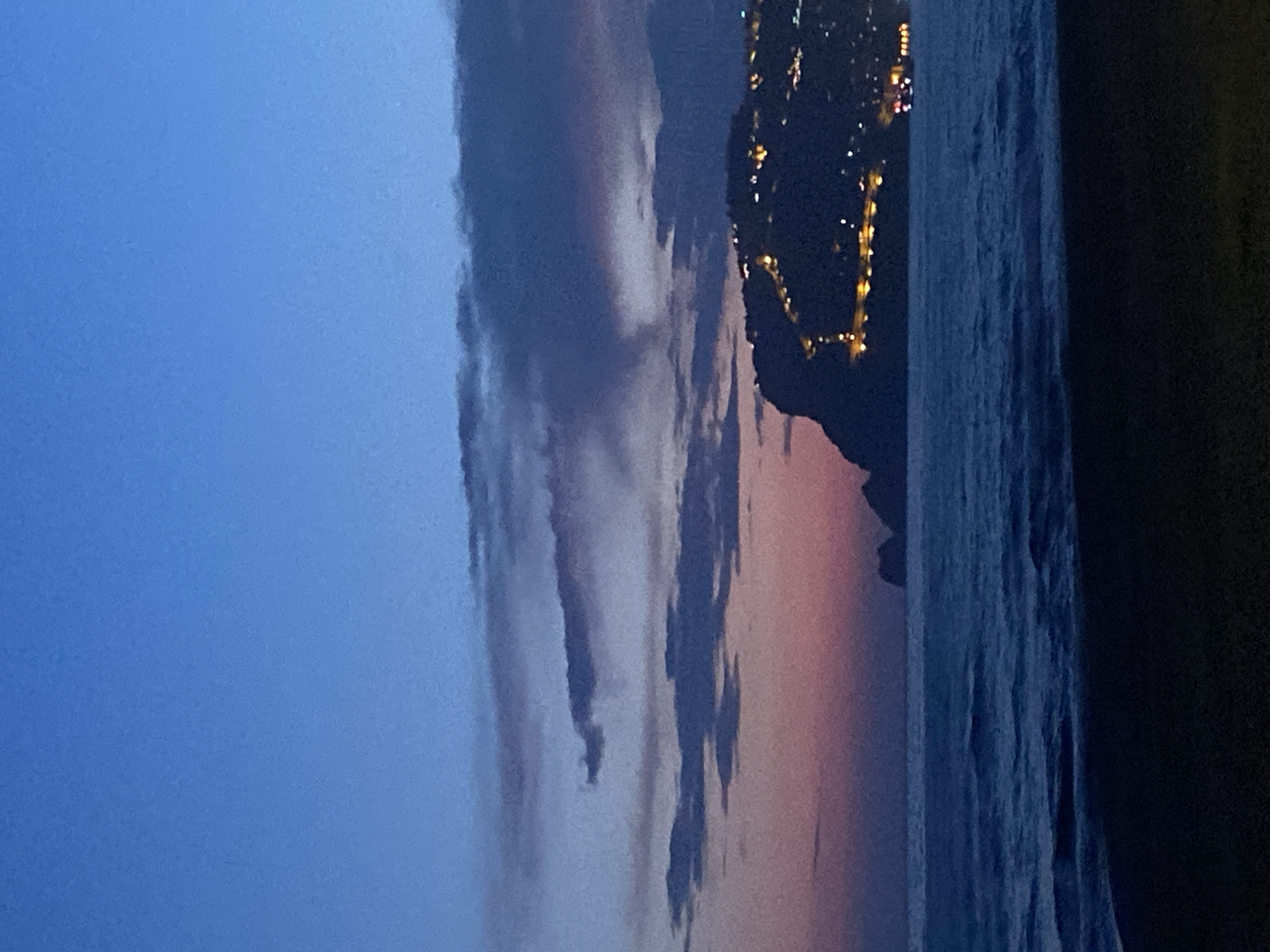 The image shows a nighttime view in Alanya, Turkey. There is a view of the sea, with the city lights twinkling on the horizon. Clouds are visible in the sky, with a gradient of colors ranging from blue to pink as the sunset fades. A dark landmass with city lights is discernible on the right, and the dark color of the sea.