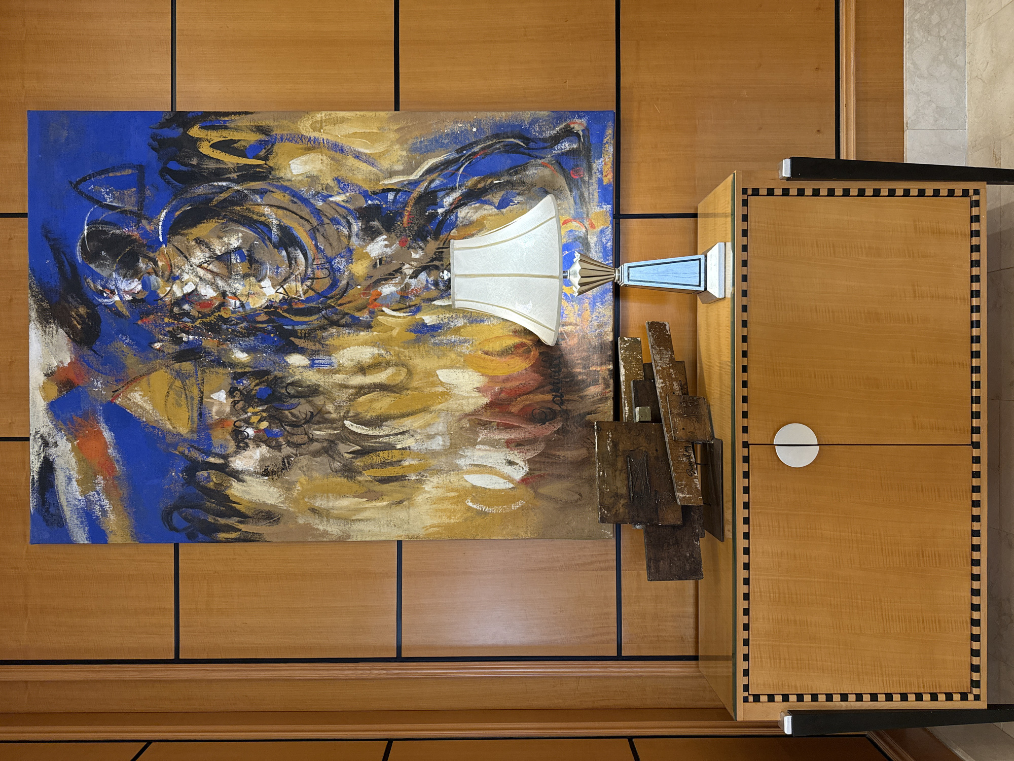 The image depicts an interior scene featuring a wooden wall paneling with black trim, a large abstract painting with bold colors of blue, yellow, red, and white, and a contemporary wooden cabinet. On top of the cabinet sits a table lamp with a white lampshade and a decorative base, and a small abstract sculpture made of layered metal pieces. The cabinet itself is made of light-colored wood with a glass top and features two doors with a single round white handle in the center. The cabinet is adorned with a black and white checkered border along the top and bottom edges. The overall aesthetic is modern and artistic. The location is identified as Sharm El Sheikh, Egypt.