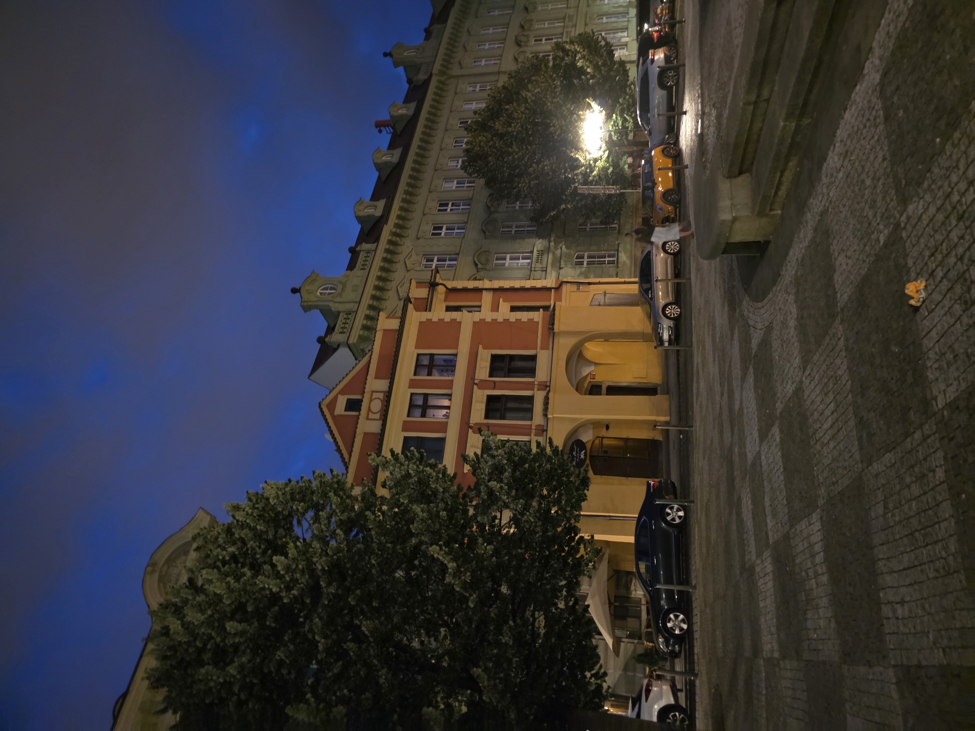 In an urban setting at dusk, a street scene unfolds with several buildings lining a cobblestone plaza. The architecture is varied, with a prominent red-and-yellow building featuring arched doorways and a lighter green building with numerous windows extending further down the street. Large trees with dense foliage are interspersed, providing shade and visual interest. Several cars are parked along the street, including a dark sedan, a silver hatchback, an orange mini cooper, and a light-colored SUV. The sky is a deep blue, transitioning from twilight, with wispy clouds visible. The ground is paved with large, dark cobblestones arranged in a checkerboard pattern in some areas and radiating patterns in others. A low stone structure with steps is visible in the foreground. The overall atmosphere suggests a quiet evening in a historic European city.