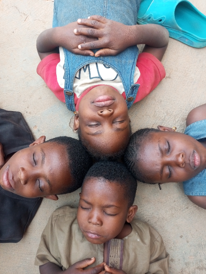 A high-angle photograph shows four individuals, presumed to be children of West African descent, lying supine on a light brown, uniform surface, possibly concrete or packed earth, in Tempure, Nigeria. Their heads are centered and closely arranged in a cruciform pattern. All four subjects have their eyes closed.
The uppermost subject wears blue denim overalls over a red and white printed shirt, with hands clasped on the torso. A single light blue slide sandal is positioned to the upper right of this individual. The subject on the left wears a dark-colored top. The subject on the right wears a blue denim garment. The lowermost subject wears a light brown tunic with dark embroidered vertical stripes on the chest, with hands clasped over the torso. No other objects or environmental features are clearly visible.