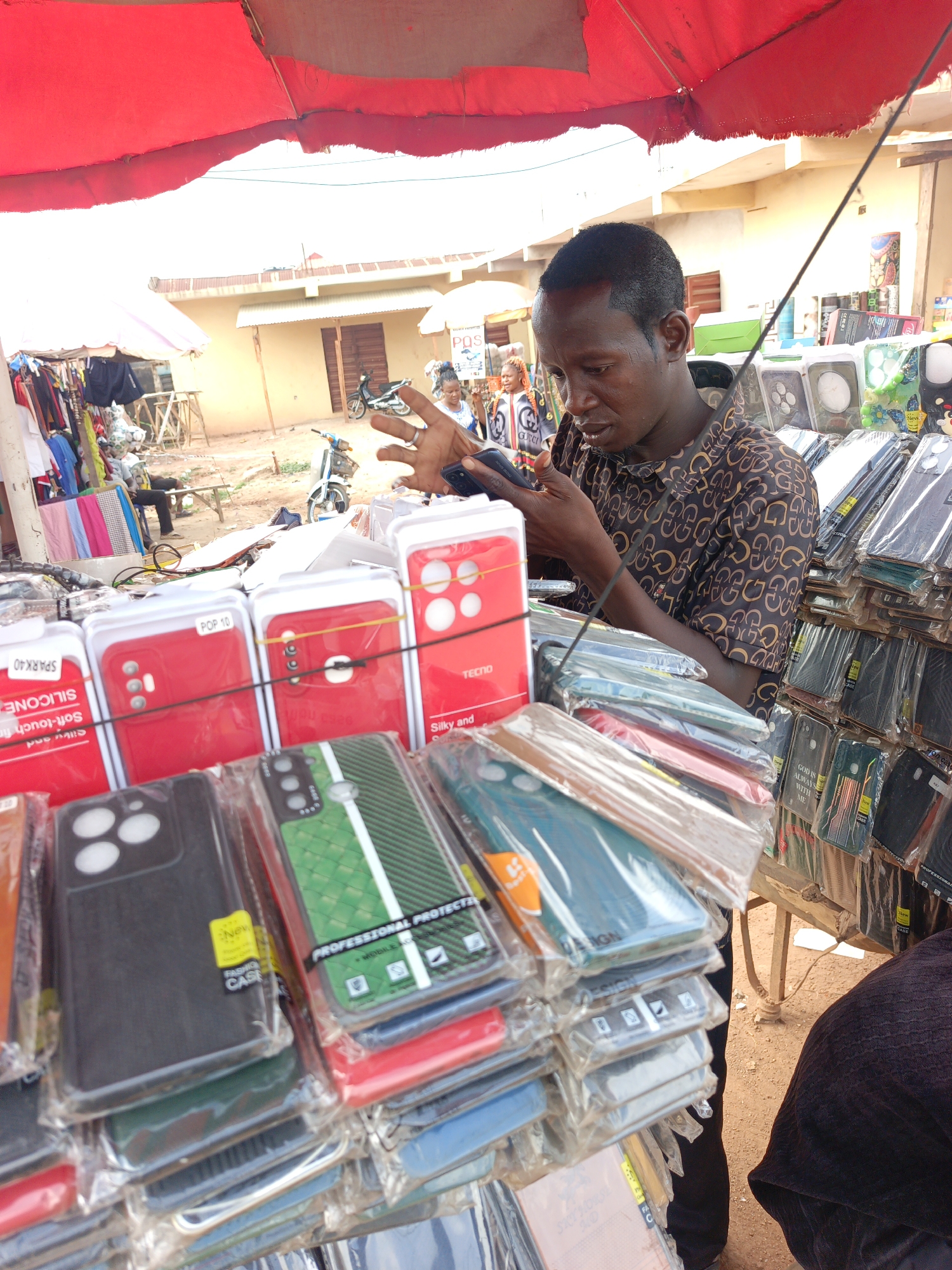 A man in Akwanga, Nigeria, stands at an outdoor stall under a red canopy, engrossed in his mobile phone. He is surrounded by a diverse collection of packaged phone cases, stacked on a table, with visible brands such as Tecno. The scene shows a commercial environment, likely a market, with buildings, other vendors, and bicycles visible in the background. The cases vary in design and features, including cases labeled "PROFESSIONAL PROTECTION," "FASHION CASE," and cases with the word "SILICONE" visible. One case features the words "GOD IS ALWAYS WITH ME." The man's clothing is a patterned brown shirt. Other people and activities are visible in the background, contributing to a bustling outdoor market setting.