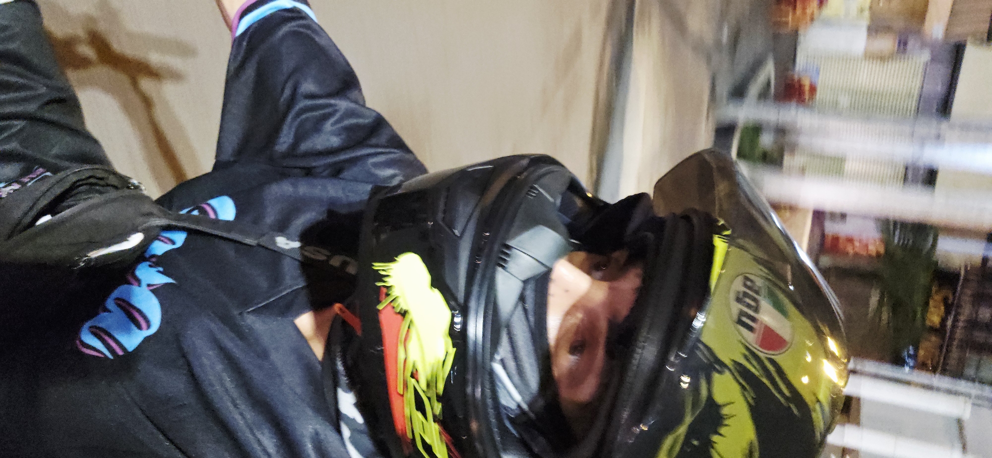 A person wearing a black and neon yellow AGV motorcycle helmet and a black jacket with blue and purple stylized figures is visible in the photograph. The person's face is visible through the helmet's visor. A cross-body bag is on their shoulder. The background shows a blurred urban scene at night, consistent with city street lighting. Based on the photograph's metadata, the image was taken in Nha Trang, Vietnam.