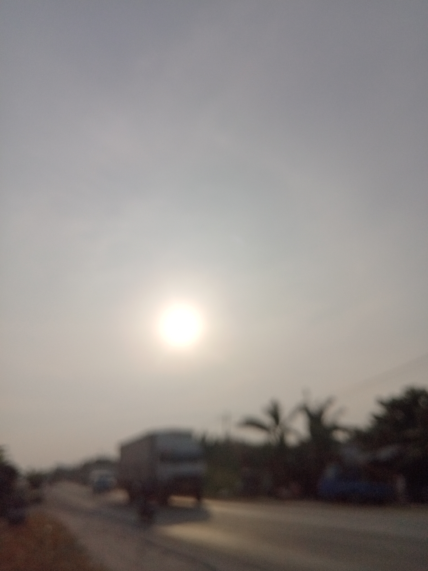 The image depicts a street scene in Kubang Jaya, Indonesia, under hazy sunlight. A large, dark truck is prominently visible on the right side of a road, moving away from the viewer. To the left of the truck, another vehicle, possibly a car, is also on the road. The road itself appears to be paved. Along the right side of the road, silhouetted trees, including palm trees, create a natural boundary. The sky is overcast with a bright, diffused sun visible towards the upper left quadrant of the image, suggesting it is either sunrise or sunset. The overall atmosphere is indistinct due to the low resolution and blurriness of the image.