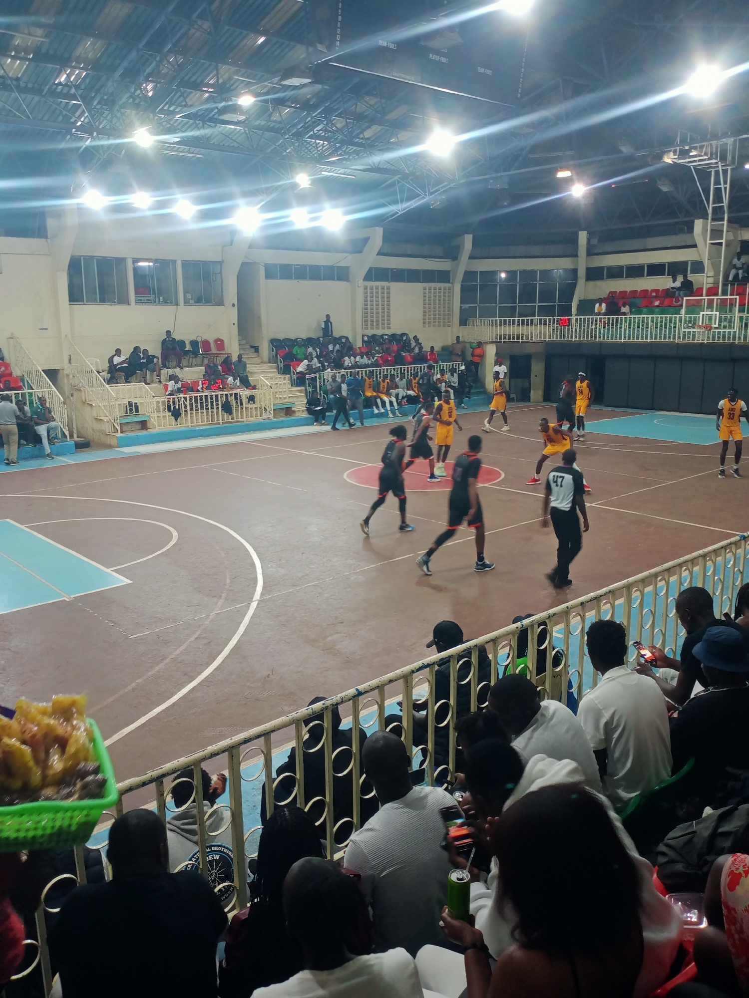 The image shows a basketball game being played indoors in a stadium in Nairobi, Kenya. Spectators are watching from the lower level, behind a railing. Some are holding phones and other objects. The basketball court has a brown surface with light blue areas around the perimeter. Two teams are actively playing. One team is wearing black and red uniforms while the other team is wearing yellow and black uniforms. A referee wearing a black and white striped shirt and black pants is on the court. More spectators are seated in the upper level of the stadium. Stadium lights illuminate the scene.