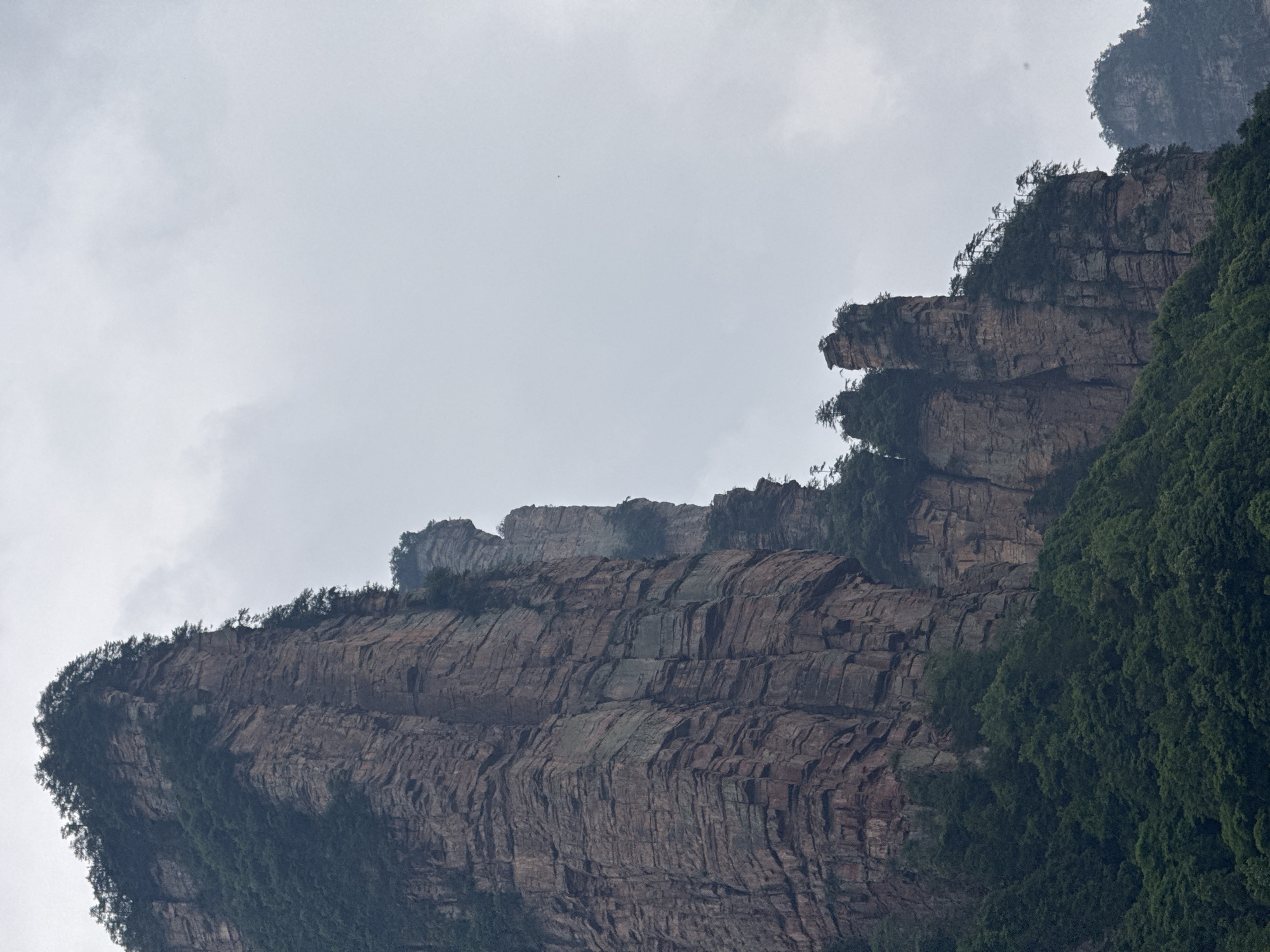 The image depicts a mountain landscape in Zhonghu, China. The mountains are characterized by steep, layered rock formations. Green vegetation covers parts of the mountains, particularly on the right side of the frame. The sky is overcast with grey clouds.