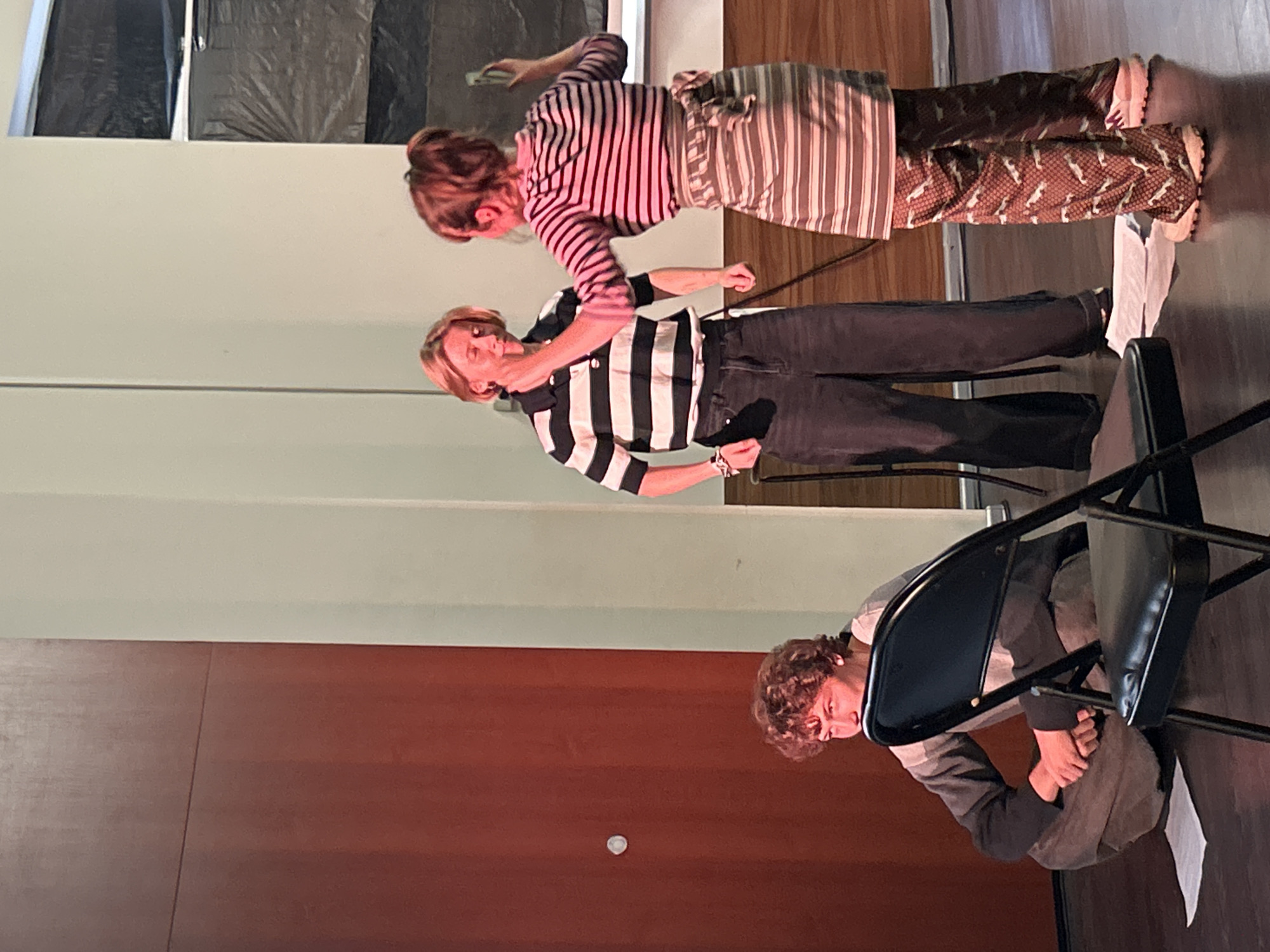 In an indoor setting with wooden flooring and pale green walls, three individuals are present. On the left, a person with curly brown hair is crouched on the floor, leaning forward, with their hands resting on their knees. In the center, a person with short blonde hair wears a horizontally striped black and white polo shirt and dark, wide-leg trousers. They are standing, facing right, with their left hand touching their face. To the right, a person with red hair tied in a ponytail is wearing a horizontally striped pink and white long-sleeved shirt and a striped apron over patterned trousers. They are standing, facing left, and appear to be interacting with the person in the center. A black folding chair is positioned in the foreground, partially obscuring the person on the left. There is a large, dark red wooden door on the far left of the image.