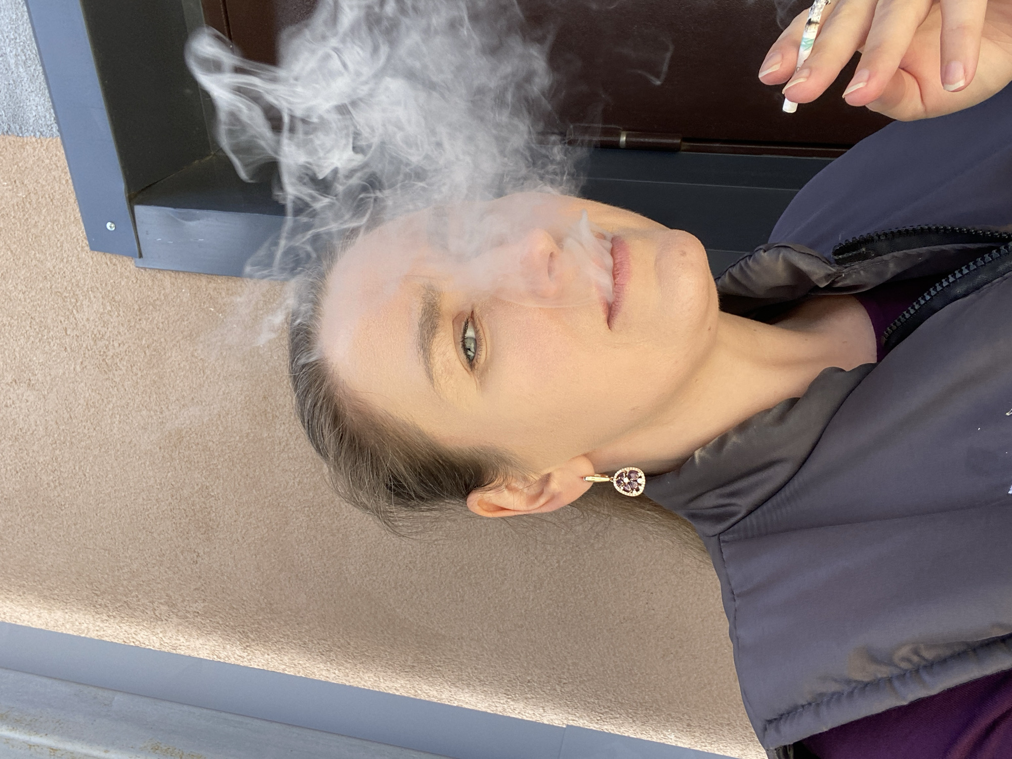A close-up, rotated portrait shot captures a woman exhaling smoke, likely from a cigarette held in her right hand, which is visible in the upper right corner. The woman's head and shoulders dominate the right side of the frame, with white smoke plumes rising from her mouth and nose, partially obscuring her face. Her eyes are open, gazing towards the viewer with a neutral, contemplative expression. She has light skin, and her hair is pulled back, with a few strands visible near her ear. A distinctive gold earring with a round, reddish-purple gem is visible on her left earlobe.
She wears a dark gray, possibly navy, puffer jacket with a visible zipper, over what appears to be a purple garment underneath. The background consists of a textured beige or light brown surface on the left and a dark gray or black structural element, possibly a window frame or door frame, running vertically behind her head. The overall lighting suggests daytime, though no harsh shadows or direct sunlight are present. The image conveys a moment of calm or introspection amidst the act of smoking. No text is visible in the image. The scene is set in Yekaterinburg, Russia.