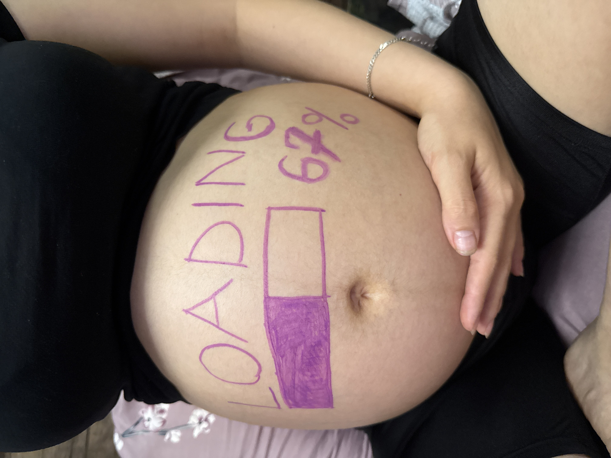 A pregnant woman's belly is the central focus of this image. The word "LOADING" is written in purple marker across the top of her belly. Below the word, a purple progress bar is drawn, filled about halfway, indicating a loading status. To the right of the progress bar, "67%" is written, further indicating the progress. The woman is wearing a black top and her hands are gently resting on her belly. The background is softly blurred, suggesting an indoor setting, possibly a home. The image does not contain any elements that would suggest a specific location in Tbilisi, Georgia.