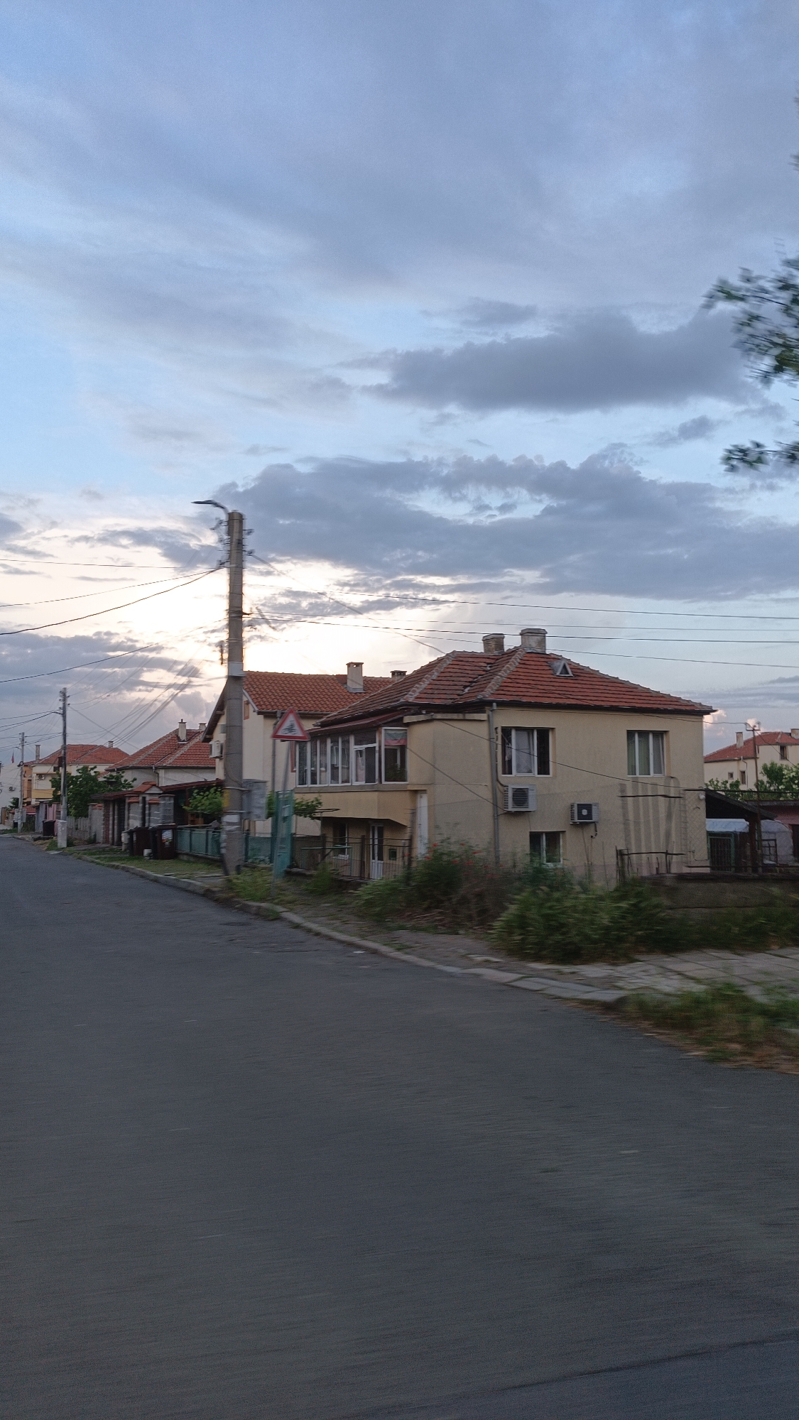 The image depicts a street in Burgas, Bulgaria, characterized by residential buildings with orange tile roofs. The structures vary in design and appearance, including visible chimneys and attached additions. A street runs alongside the buildings. An electrical pole with overhead wires is near the buildings. A road sign is also visible. The sky overhead has clouds and patches of light. Overgrown vegetation is on the curb along the road and in front of the buildings.