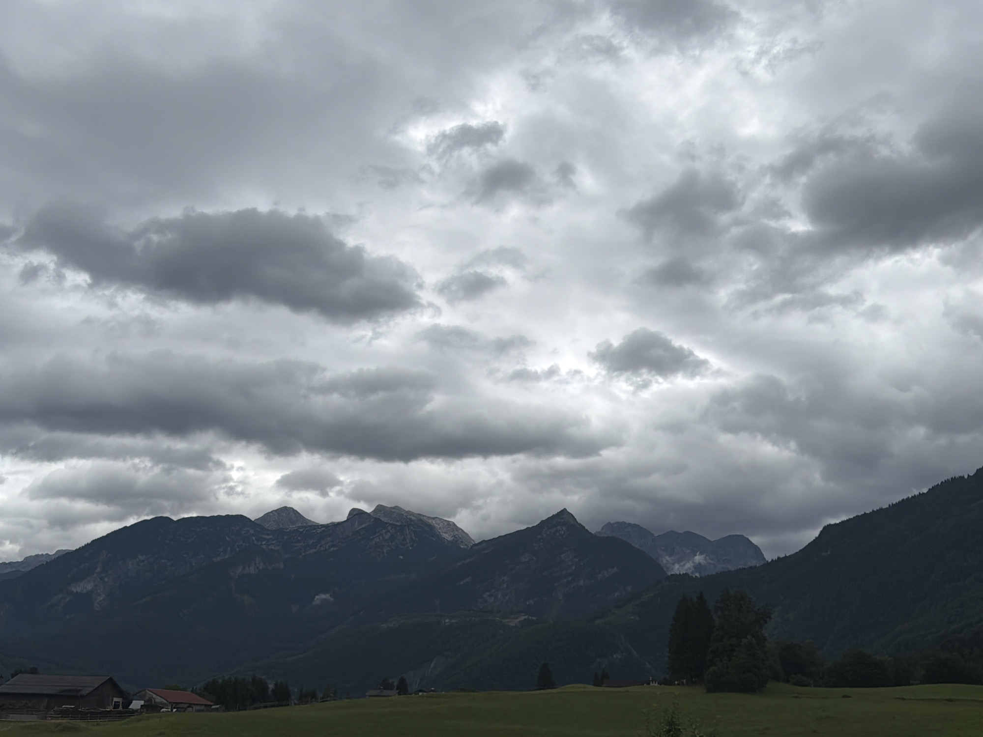 This is a medium-shot, horizontal, eye-level, color photograph of a mountainous landscape under a heavy, overcast sky. The scene depicts the region of Torren, Austria. In the foreground, a green, grassy field stretches across the bottom portion of the image. On the far left of the field, a dark brown, wooden building with a lighter brown roof is visible, partially obscured by the edge of the frame. To the right of this building, a smaller, red-roofed structure is partially visible. A few sparse trees dot the field.
The middle ground is dominated by a series of dark, imposing mountain ranges. These mountains are heavily shadowed and appear to be covered in dense foliage or rock faces. The peaks of the mountains are jagged and varied in height, with some appearing snow-capped in the distance, although this could be mist.
The background is a tumultuous sky filled with thick, gray clouds. These clouds are varied in density, with some areas appearing lighter and more diffuse, suggesting sunlight attempting to break through, while other areas are dark and ominous. The overall impression of the sky is one of impending weather, possibly rain or a storm. No people or animals are visible in the image. The environment appears to be rural and remote, with a focus on the natural landscape.