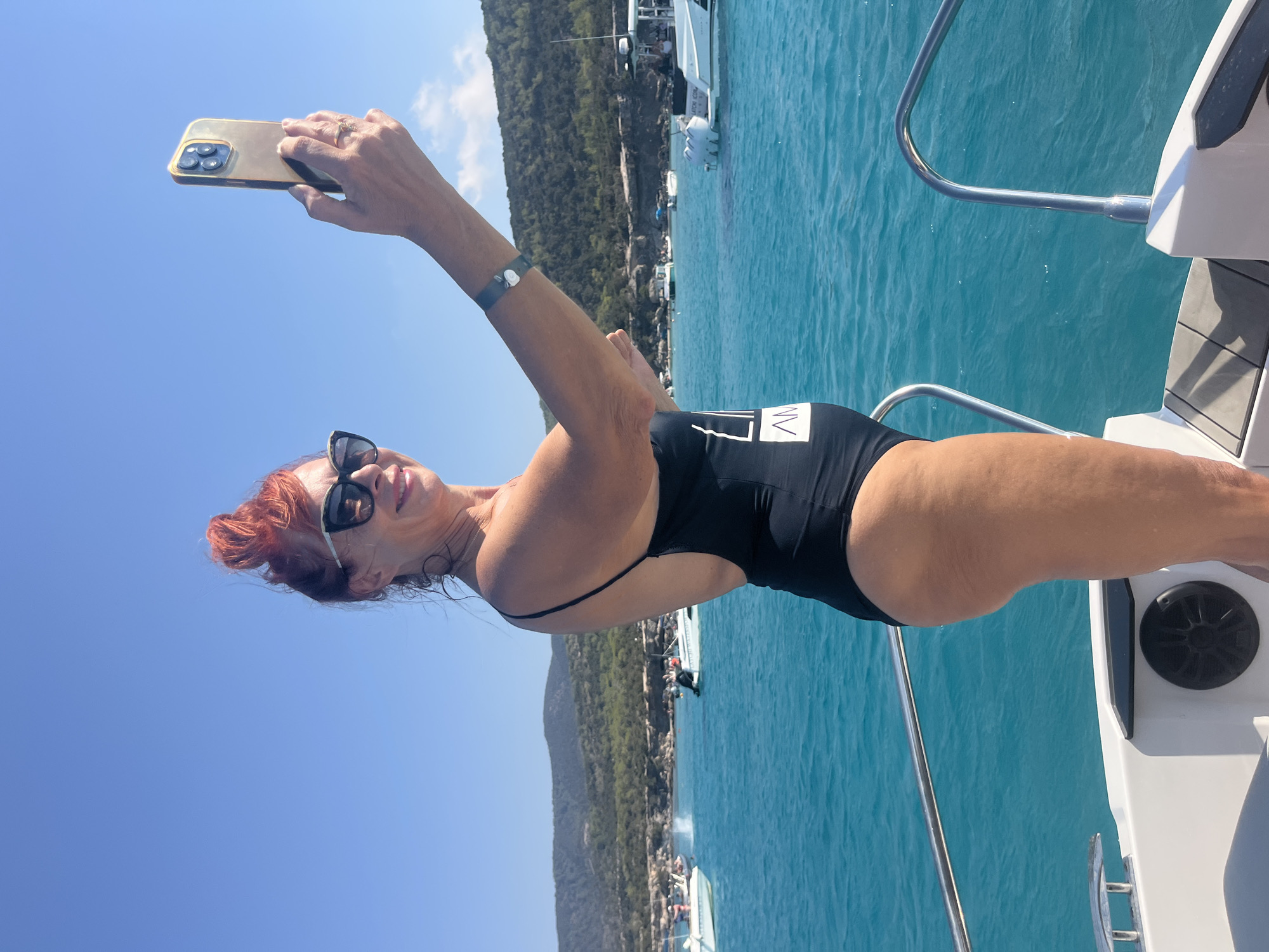 A woman with red hair styled in a bun, wearing sunglasses and a black swimsuit, stands on a boat in the bright sun. She is taking a selfie with a smartphone in her right hand. The boat is on a vibrant turquoise sea, with a coastline of green hills in the background. Several other boats are visible on the water. The scene is set in Neo Chorio, Cyprus.