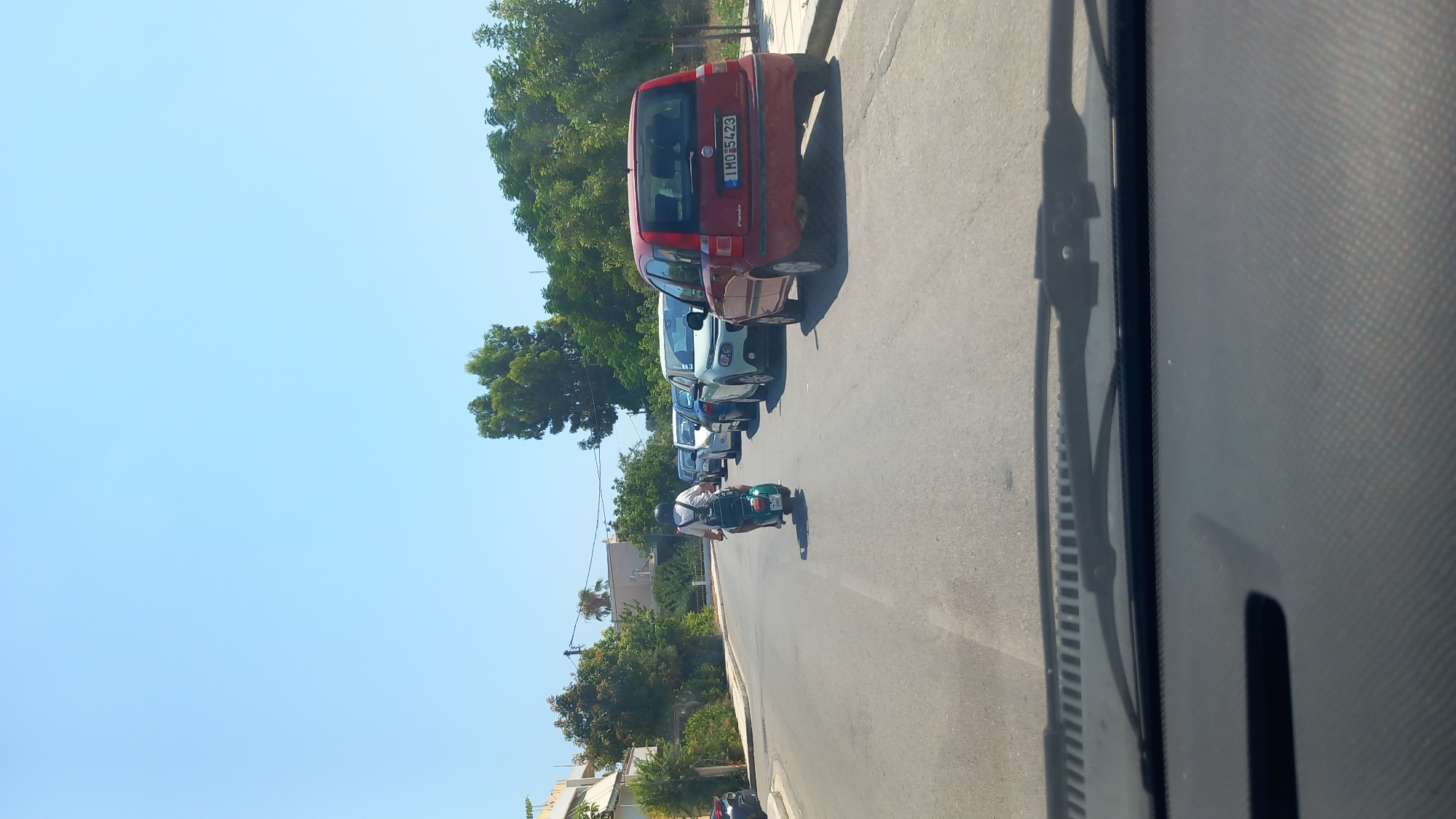 A person on a scooter drives down a street in Kos, Greece, with several cars parked on the right side of the road. The sky is clear and blue. The street is paved and appears to be in good condition. Trees and buildings line the street. The scooter is green and the person is wearing a helmet and a white shirt. The cars are various colors and models. One car in the foreground is red and has a license plate that reads "1 MO 5423". The image is taken from inside a vehicle, likely a car, as the windshield wipers are visible in the foreground.