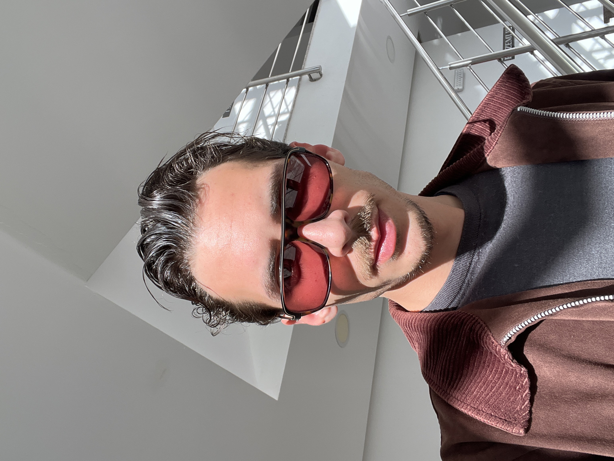 A man with short, dark, wet hair is posing for a close-up, low-angle shot. He is wearing tinted sunglasses, a gray long-sleeved shirt, and a brown corduroy jacket. The background consists of white walls and a metal staircase railing, suggesting an indoor location. The photograph appears to have been taken near or in the city of Rafailovići, Montenegro, based on contextual cues. The lighting is bright, with strong sunlight creating shadows across the scene.