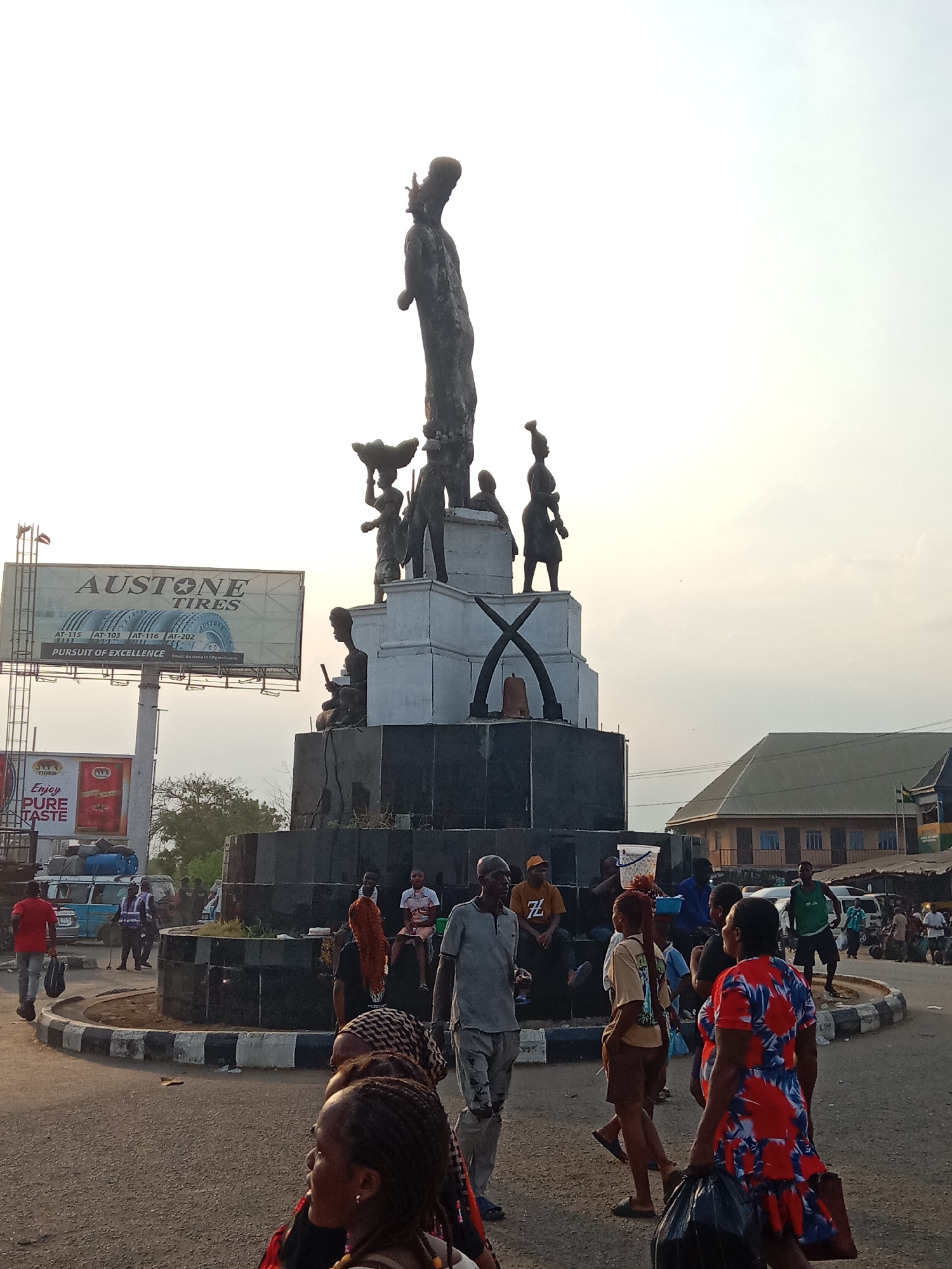 The image captures a bustling scene at the Denis Memorial Grammar School (DMGS) Roundabout in Onitsha, Nigeria. The focal point is the roundabout's monument, a tall sculpture composed of several figures mounted on a stepped, circular base with a prominent white section featuring elephant tusks. An "AUSTONE TIRES" billboard stands nearby. Numerous pedestrians are visible, their clothing and actions indicative of a marketplace or busy commercial area. The weather appears overcast, casting a somewhat muted light over the scene. Buildings and vehicles in the background complete the urban environment.
