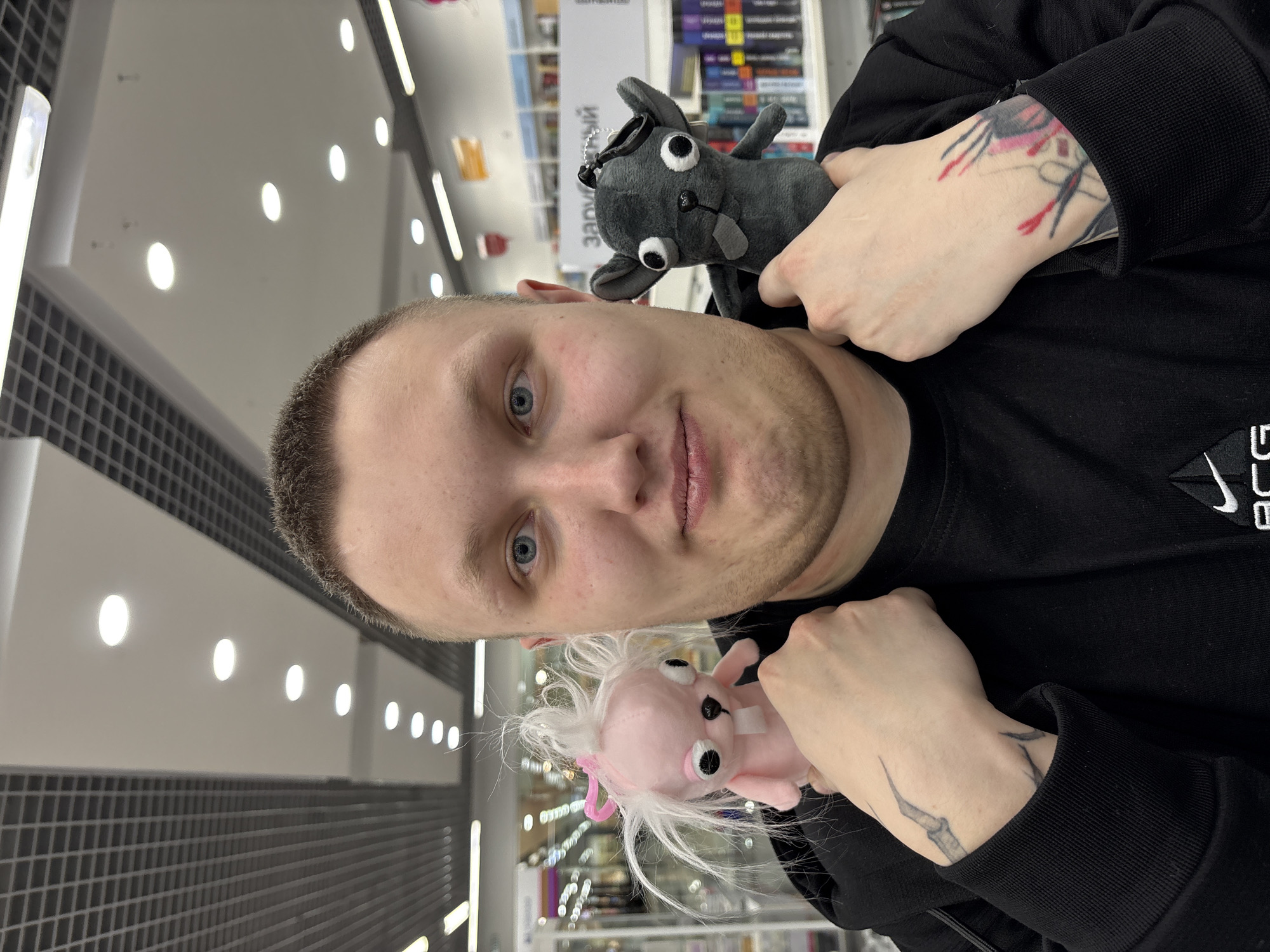A man poses for a selfie in a store in Izhevsk, Russia. He holds a gray stuffed animal on one shoulder and a pink one on the other. The man has fair skin, light brown hair, and blue eyes. He wears a black hooded sweatshirt. His hands and wrists are visible, with tattoos on both. The store has bright overhead lighting and displays shelves filled with merchandise. A wall with a dark gray tile pattern is visible in the background.