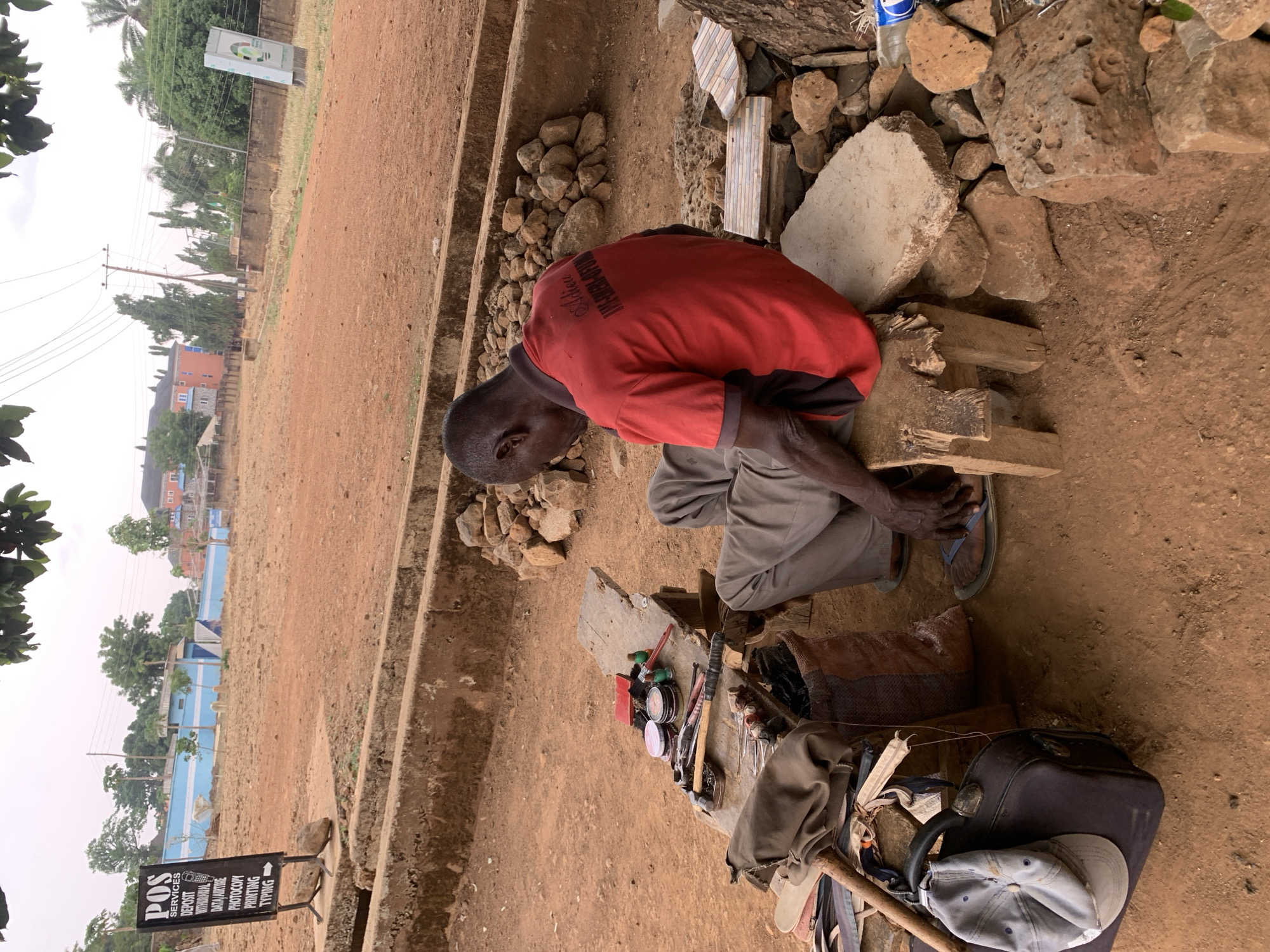 A man in a red shirt and gray pants is hunched over a makeshift workstation in Maigizo, Nigeria. He is sitting on rough wooden supports amidst scattered rocks and a concrete block. His workstation holds an assortment of tools, suggesting he may be a craftsman or repairman. Nearby, a woven bag, a piece of luggage, and a cap are placed on the ground. In the background, there's a sign advertising "POS Services" including deposit, withdrawal, data/airtime, photocopy, printing, and typing. The surrounding environment appears to be a dusty, undeveloped area with glimpses of buildings and greenery.