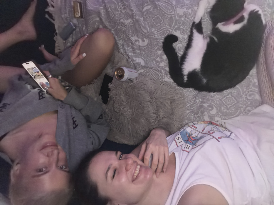 Two women lie on a patterned bedspread. One woman in a gray sweatshirt looks at a phone, while the other in a white t-shirt smiles. A black and white cat stretches out nearby. A beer can and a black phone rest on a fluffy pillow near the women. Cigarette lighter, a pack of cigarettes, and the toes of another person are also in the frame.