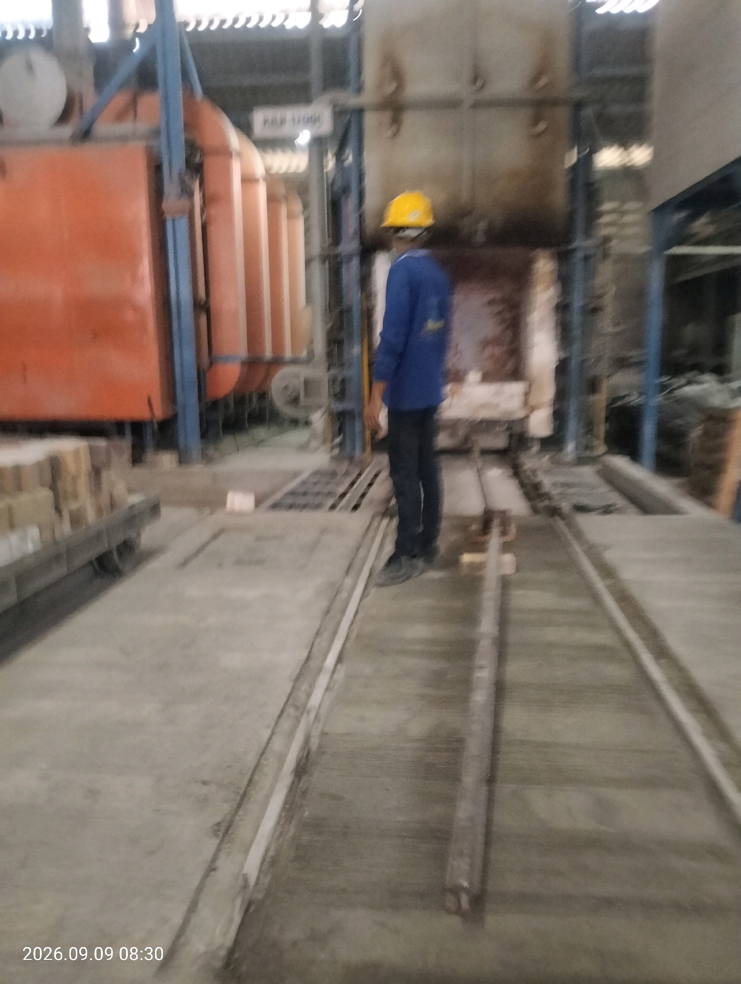 A blurred image shows a person wearing a yellow hard hat and a blue uniform in what appears to be an industrial setting, possibly a factory or processing plant. The person is standing on a concrete floor, with a large orange industrial oven or kiln visible in the background. There are also stacks of what appear to be bricks or building materials on a cart to the left. The overall scene suggests an operational work environment, likely in Bambe, Indonesia, given the timestamp in the lower left corner of the image which reads "2026.09.09 08:30".
